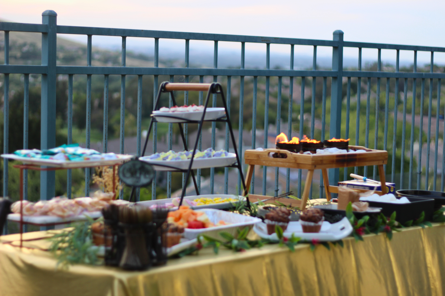 Outdoor buffet table with tiered display and chafing dishes on a balcony overlooking a scenic landscape.
