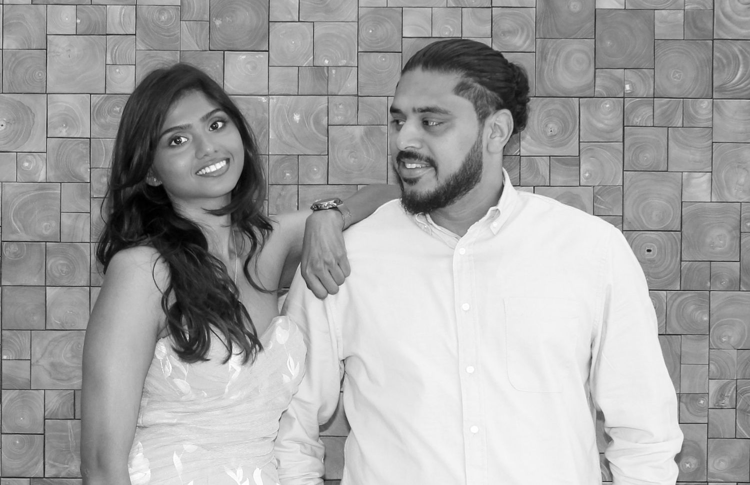 Black and white photo of a young couple posing together against a tiled wall, woman in white dress, man in white shirt.