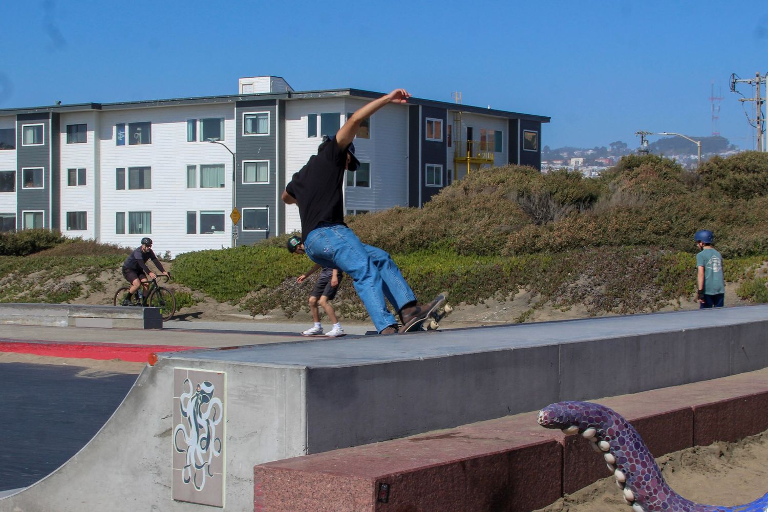 Skateboarder performing a trick at an outdoor concrete skate park near the beach with apartment buildings in the background.