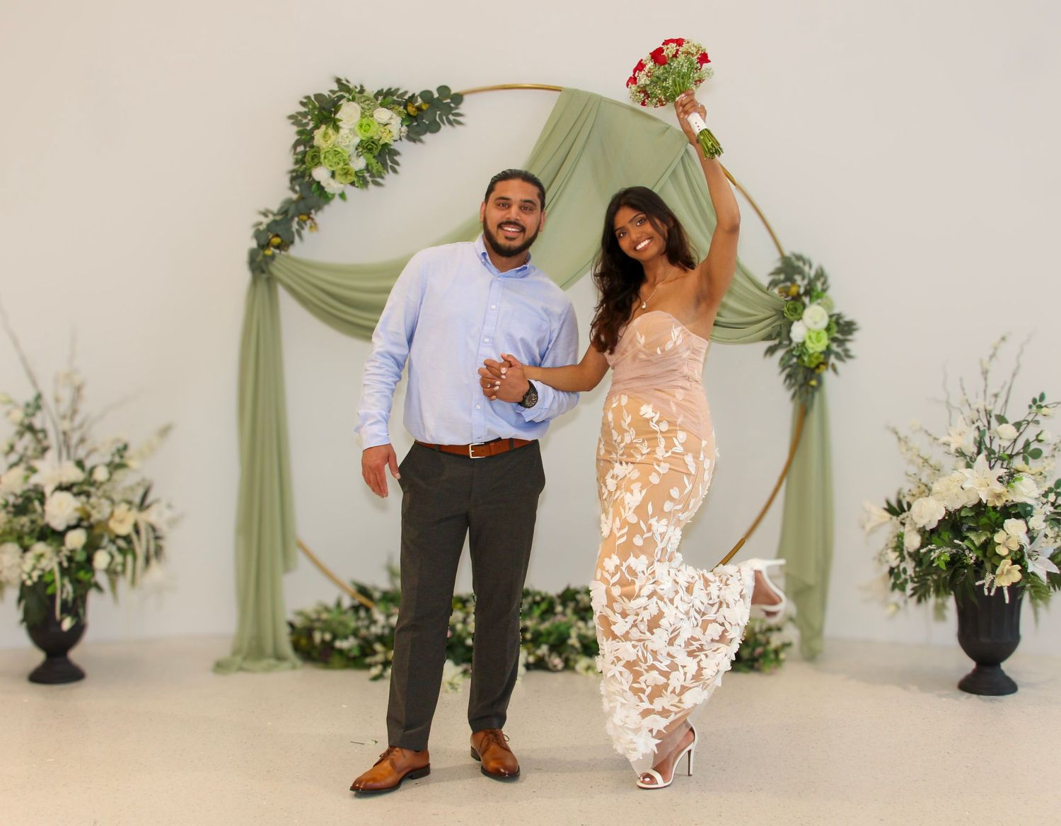 Newlyweds celebrate in front of circular floral arch, bride raising red bouquet in elegant lace gown.