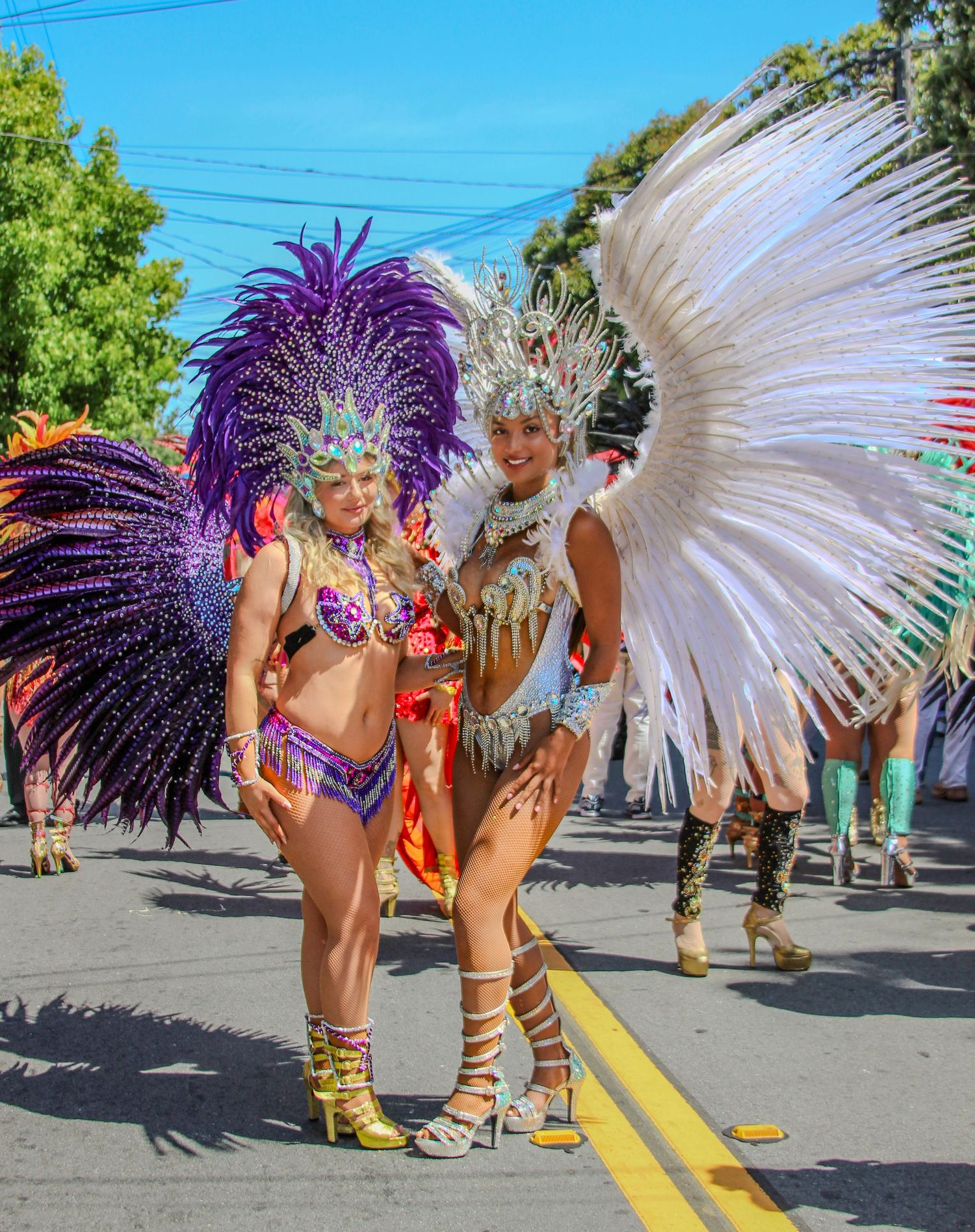 Two women in vibrant feathered carnival costumes pose on a sunny street during a colorful parade celebration.