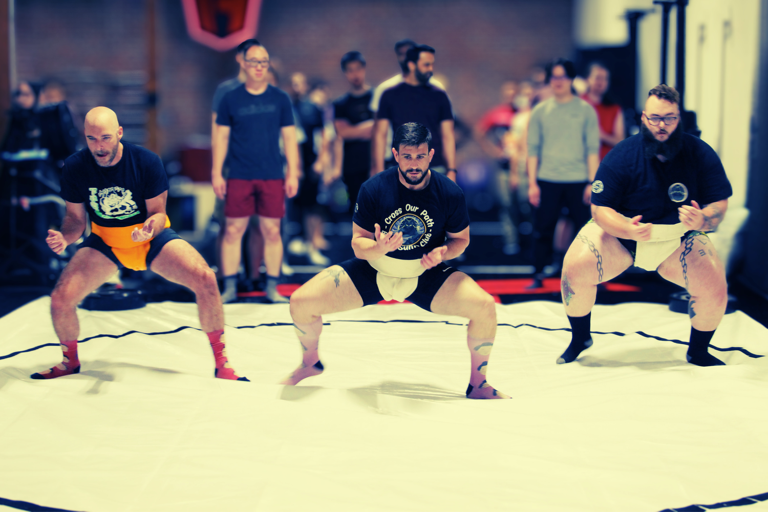Three men in sumo wrestling stances compete on a white mat during an indoor martial arts tournament.