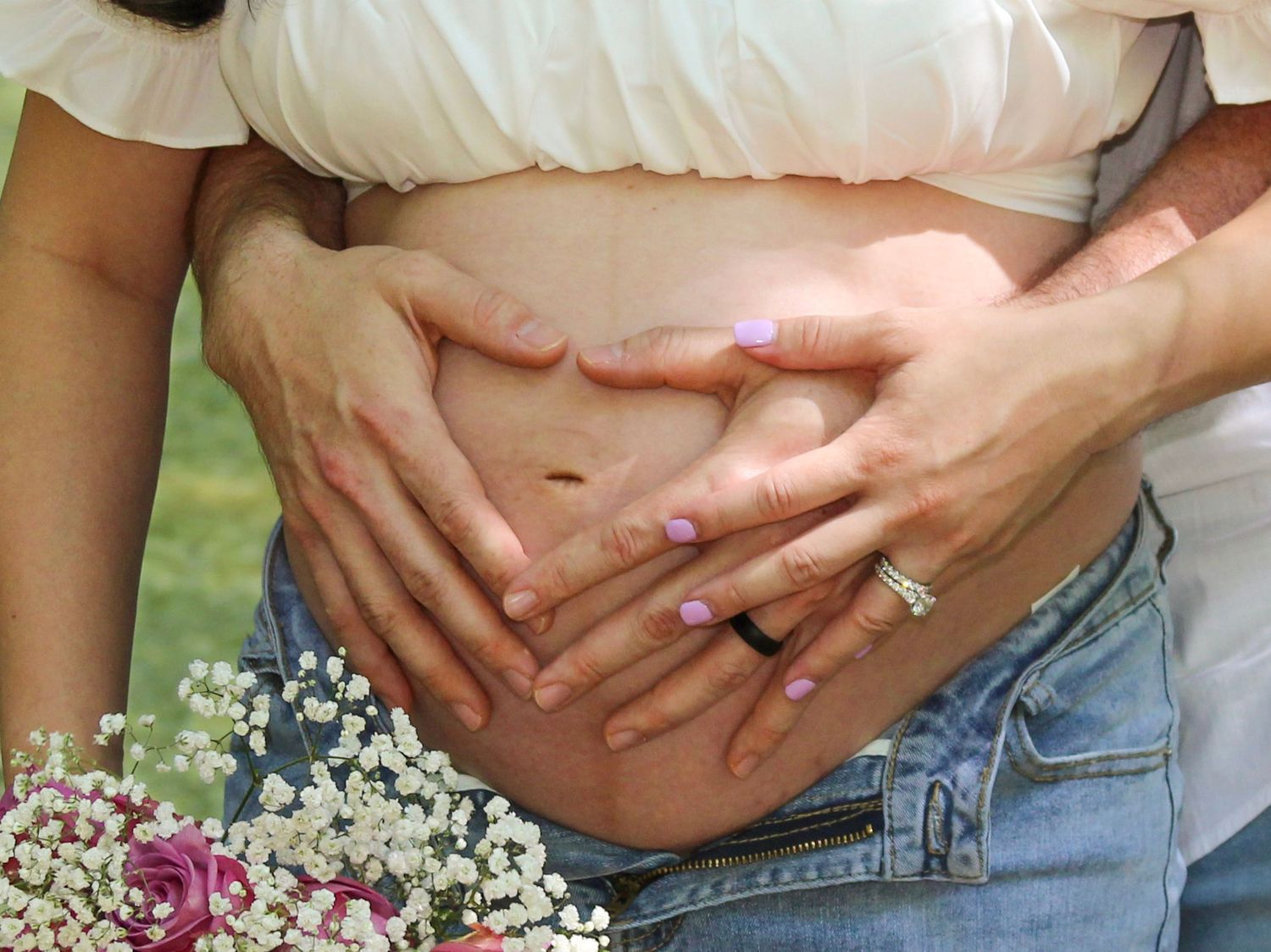 Couple forming heart shape with hands on pregnant belly, with wedding rings visible and flowers nearby.