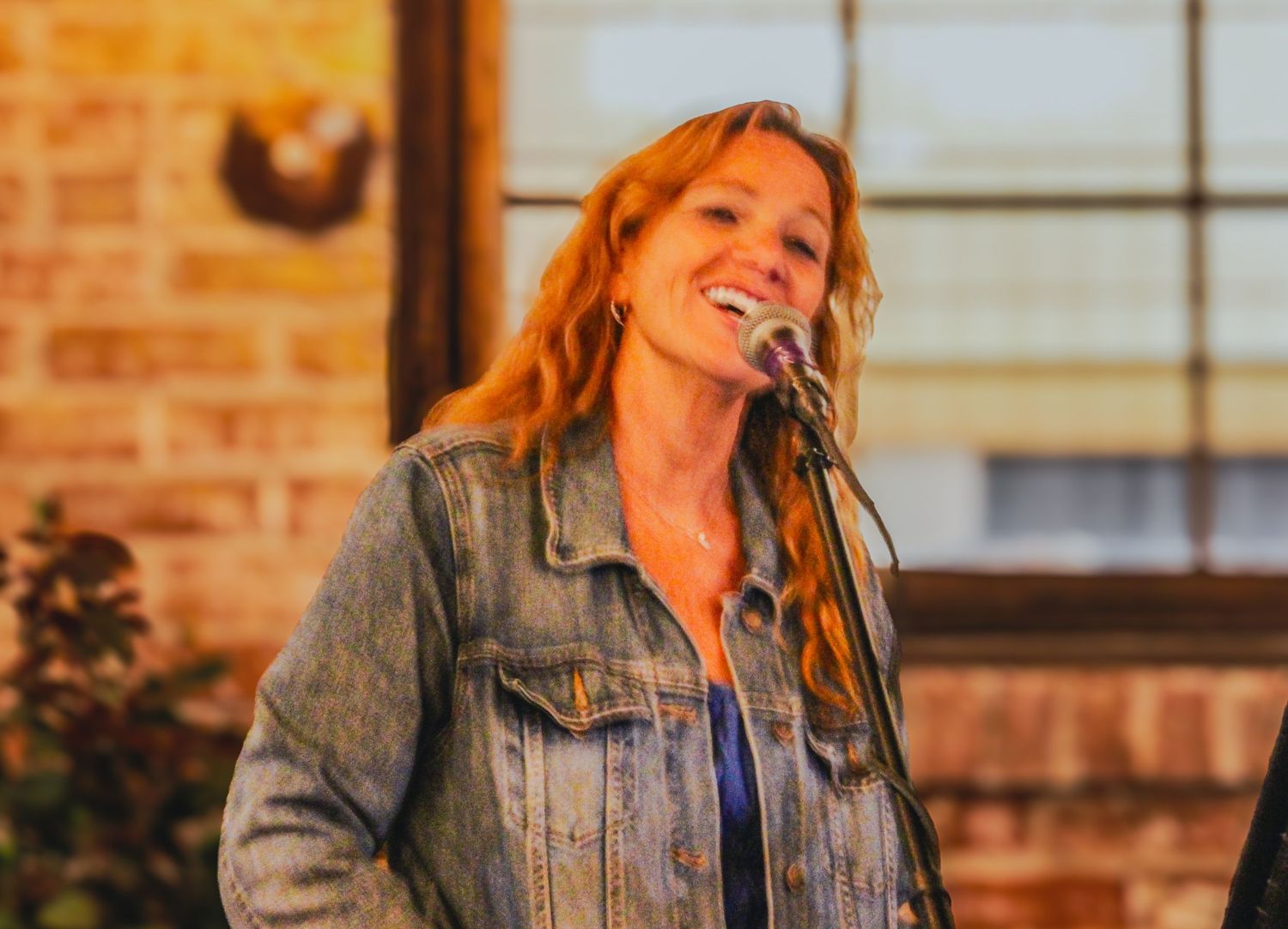 Woman with red hair singing into microphone, wearing denim jacket, performing live on stage with brick wall background.