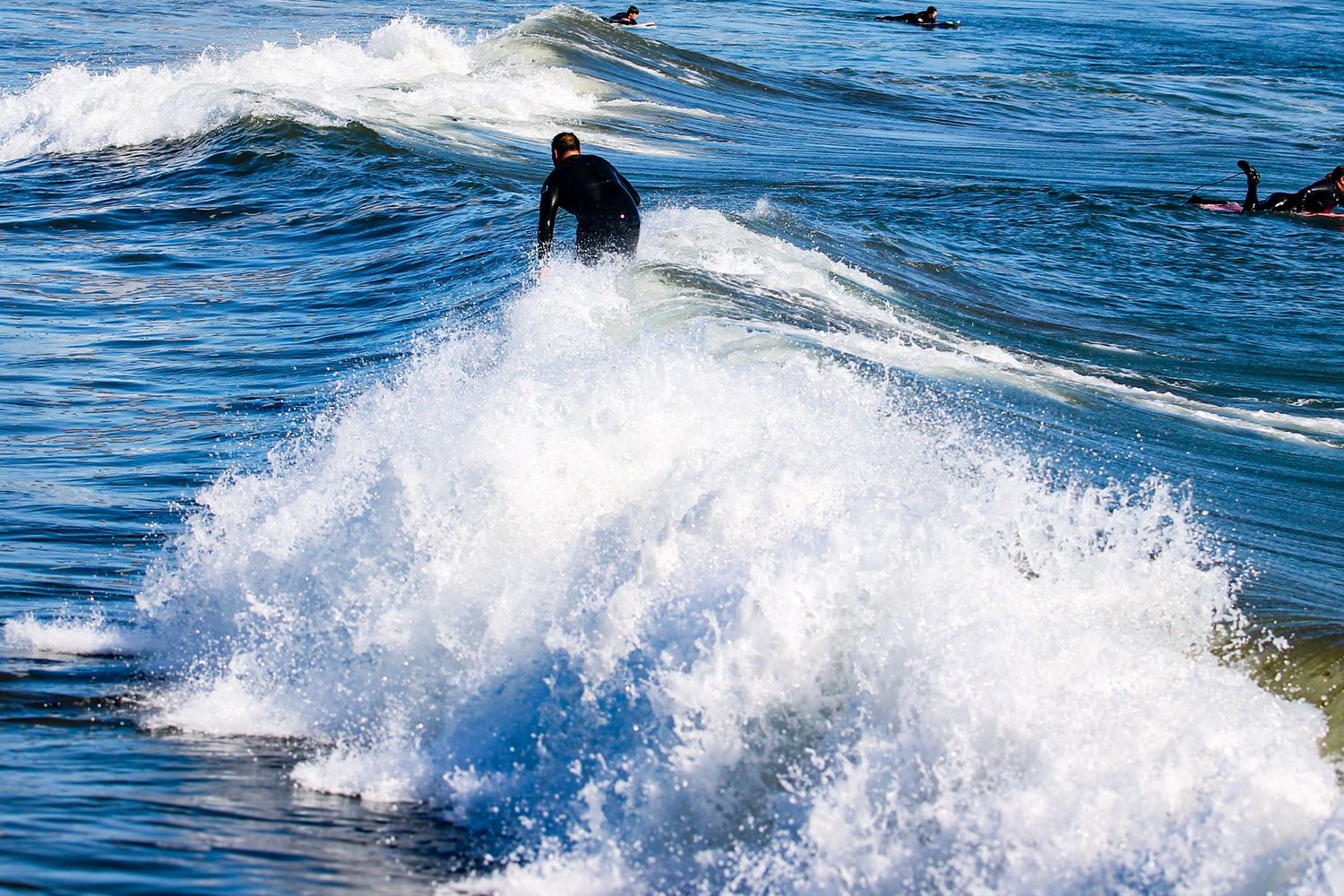 A surfer in a wetsuit rides a crashing white wave in the ocean on a sunny day.