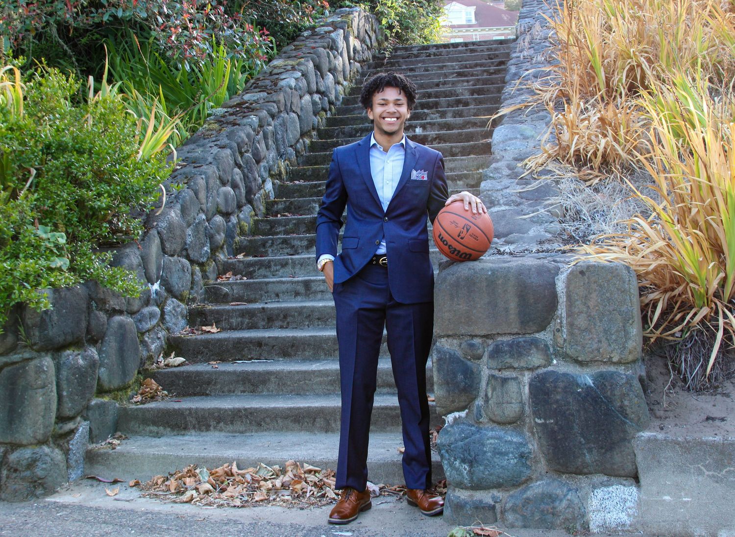 Young man in navy suit holding basketball poses confidently on stone steps outdoors.