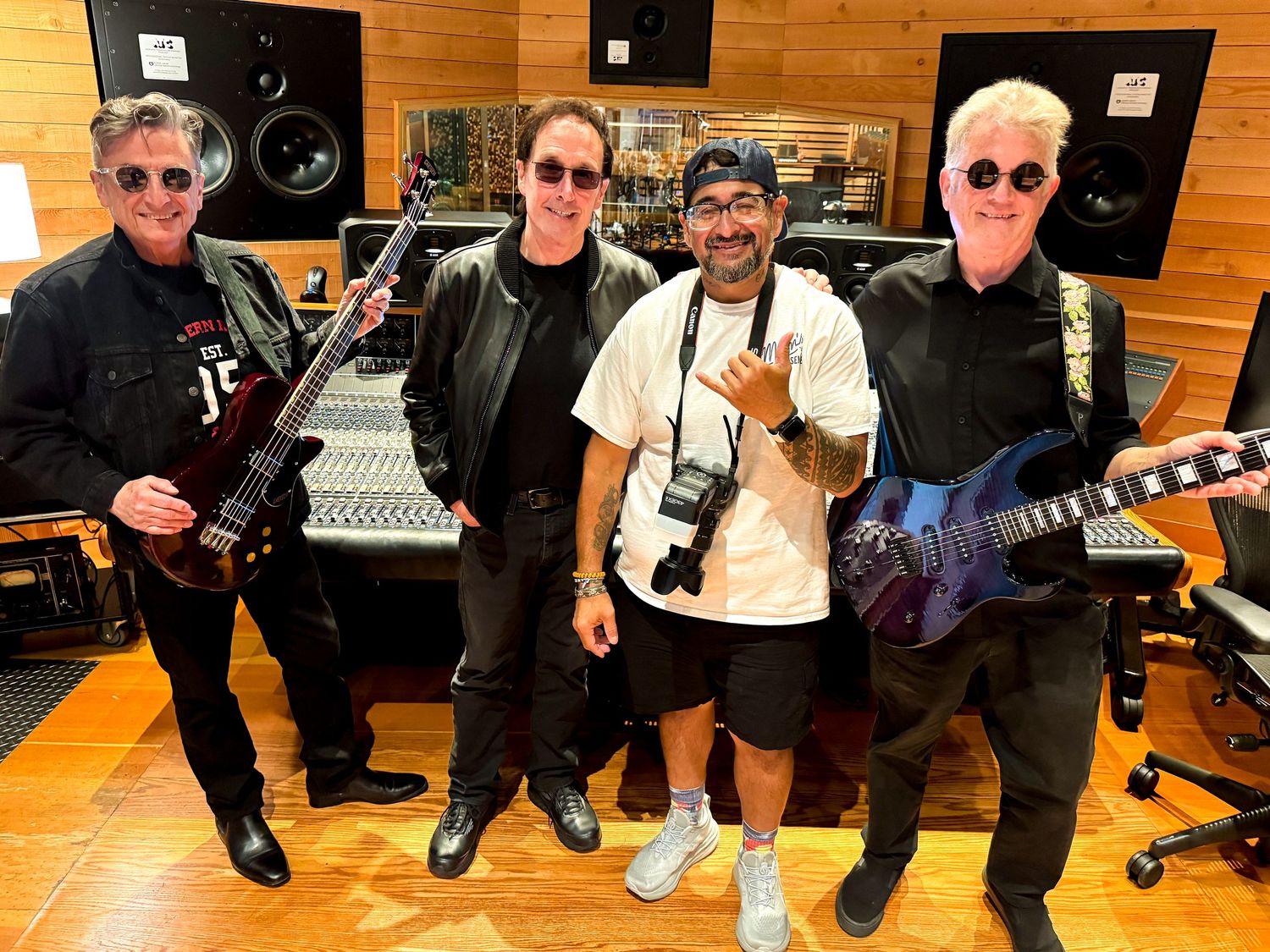 Four men posing in a recording studio, two holding electric guitars, with mixing console and studio monitors in background.