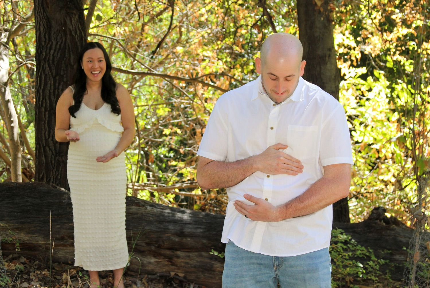 Pregnant woman in striped dress and bald man both cradling their stomachs outdoors among autumn trees.