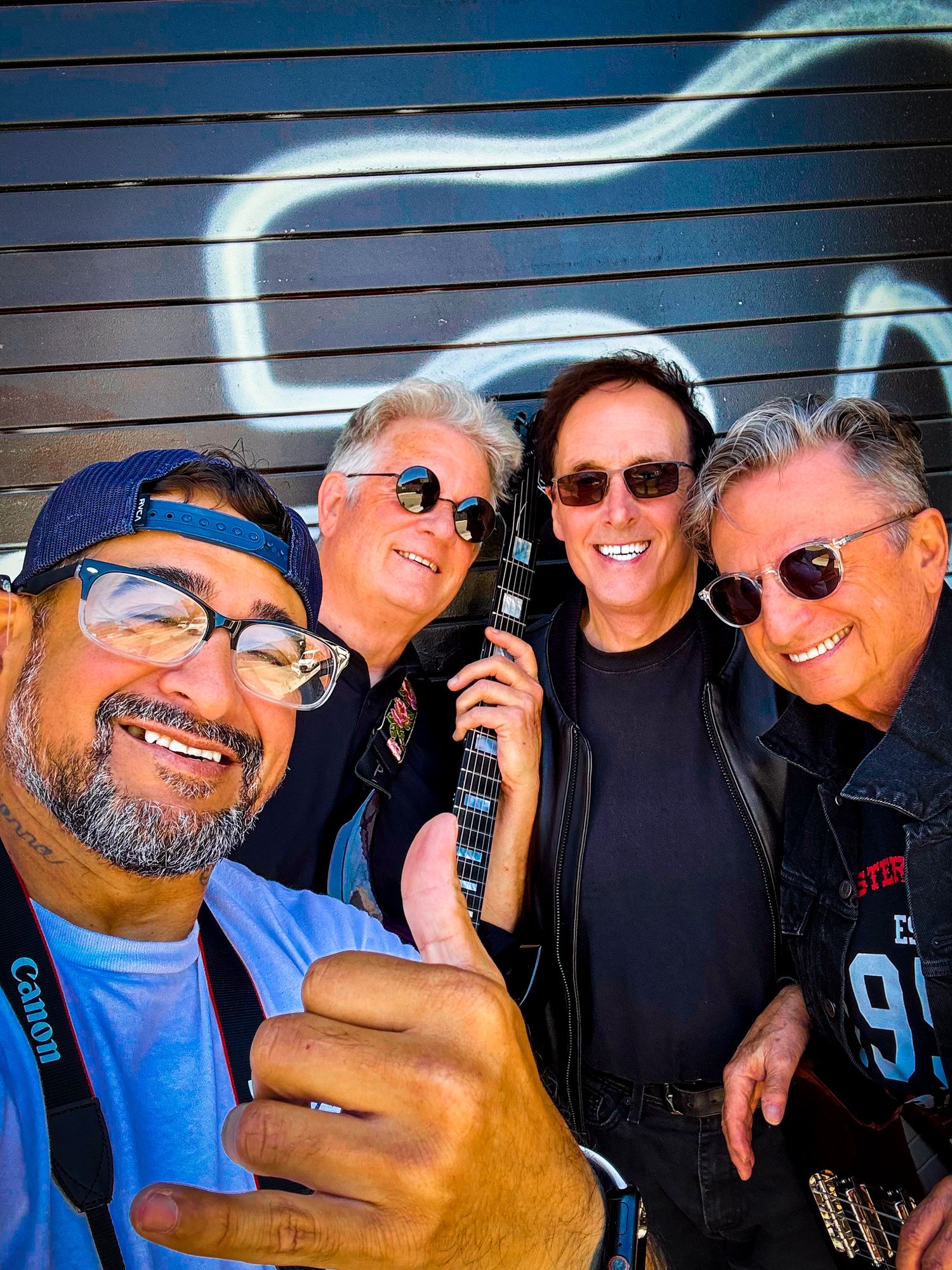 Four men smiling for a selfie outdoors, one holding a guitar, with a colorful mural backdrop.
