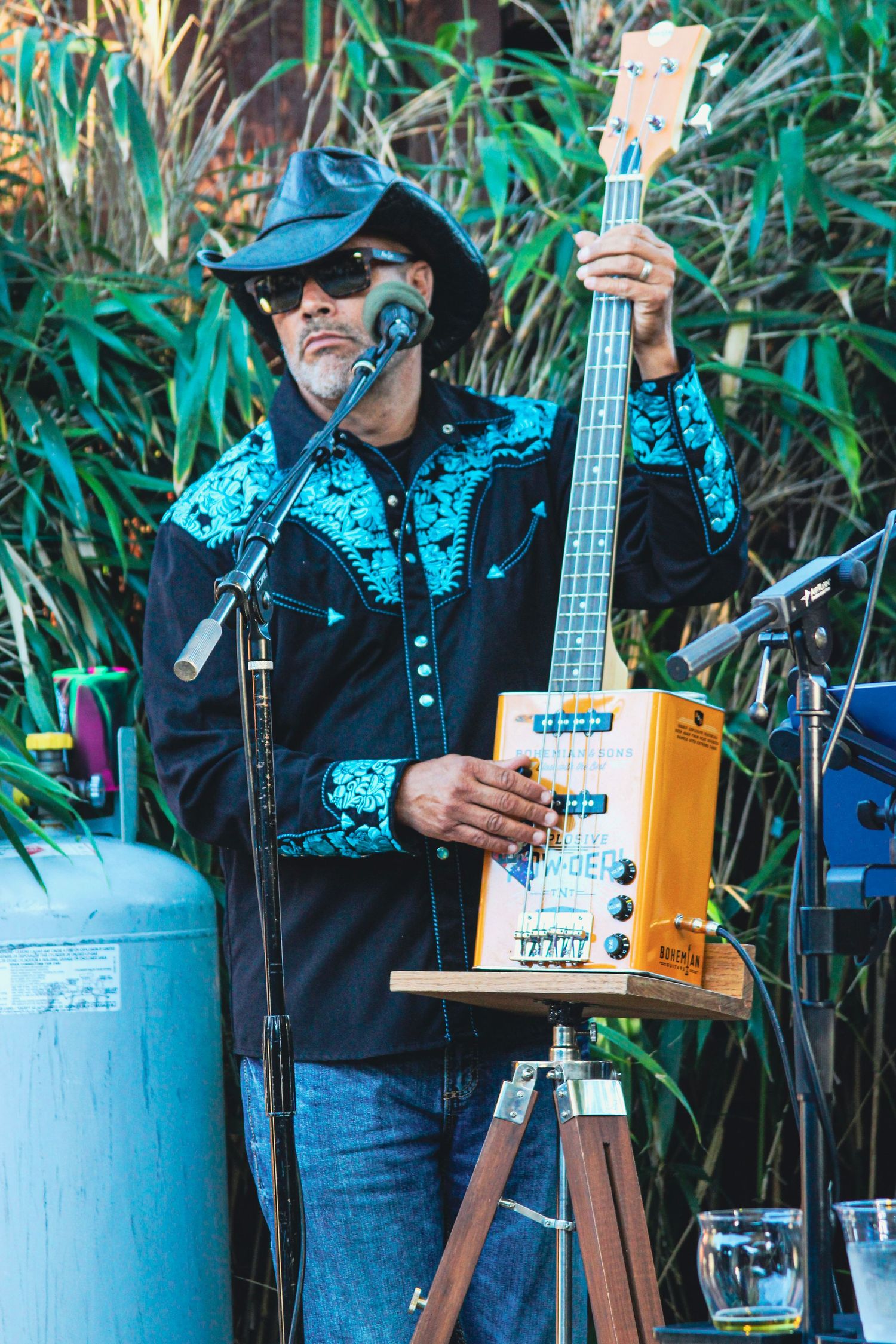 Musician in black cowboy hat and embroidered shirt plays electric bass guitar outdoors at a live performance.