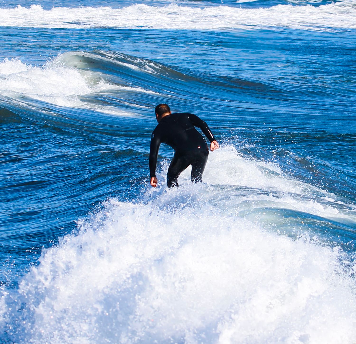 A surfer in a black wetsuit rides a wave in the ocean, surrounded by white sea foam and blue water.