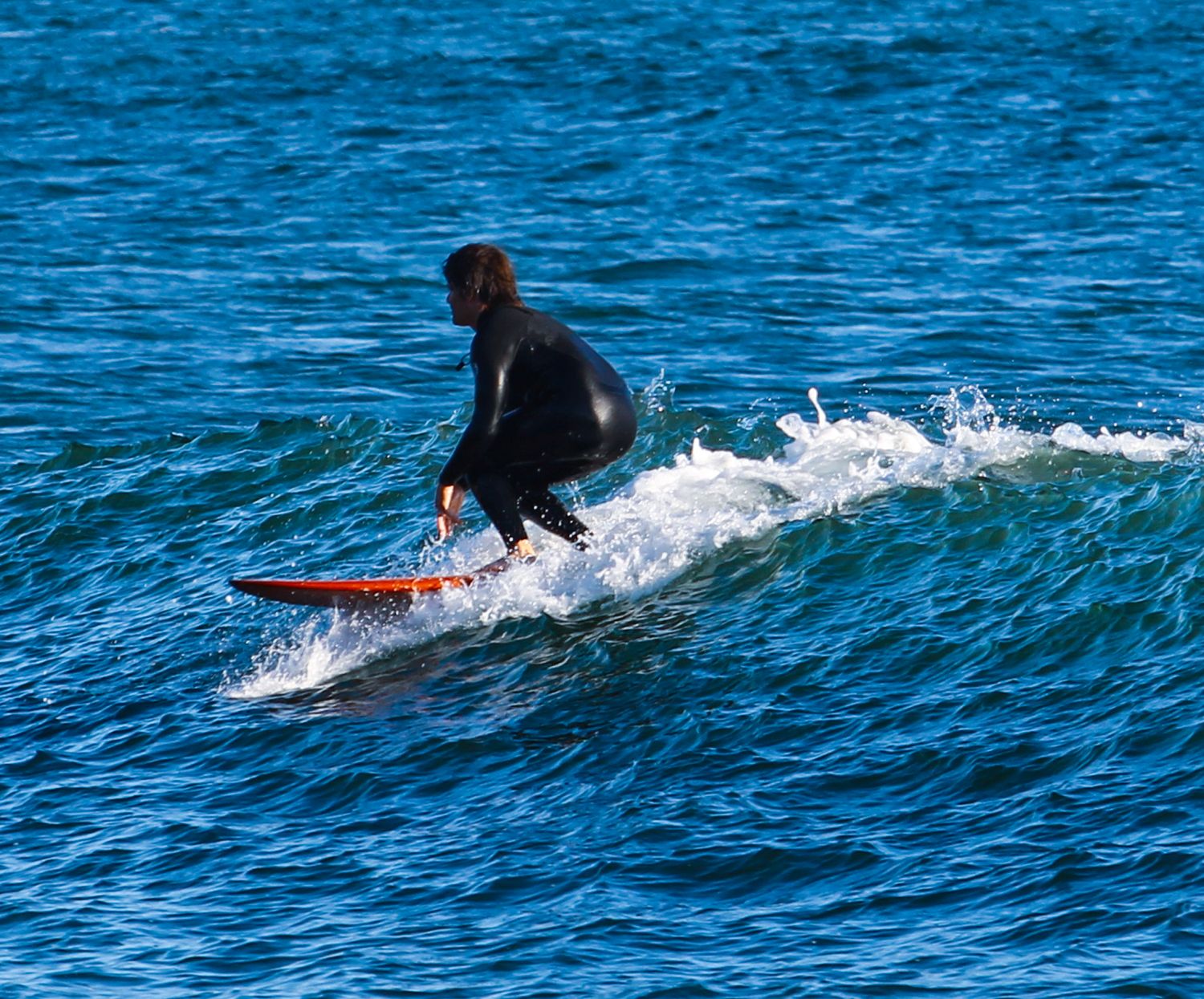 A surfer in a black wetsuit rides an orange surfboard on a blue ocean wave.