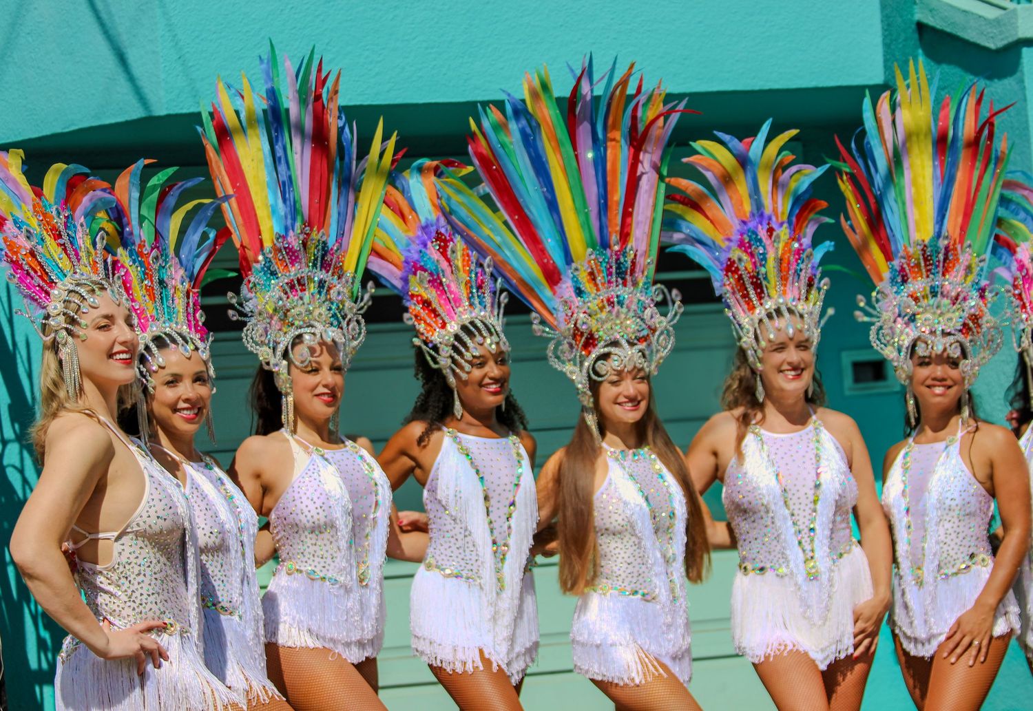 Seven women in white sequin costumes with colorful feather headdresses posing at a carnival parade.