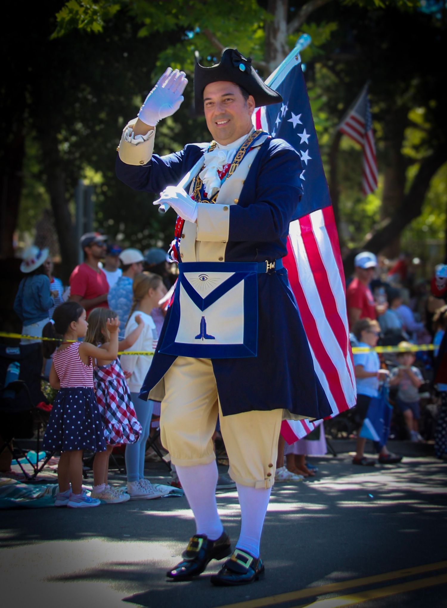 Man in colonial Freemason costume waves while carrying American flag in a 4th of July parade with crowd watching.