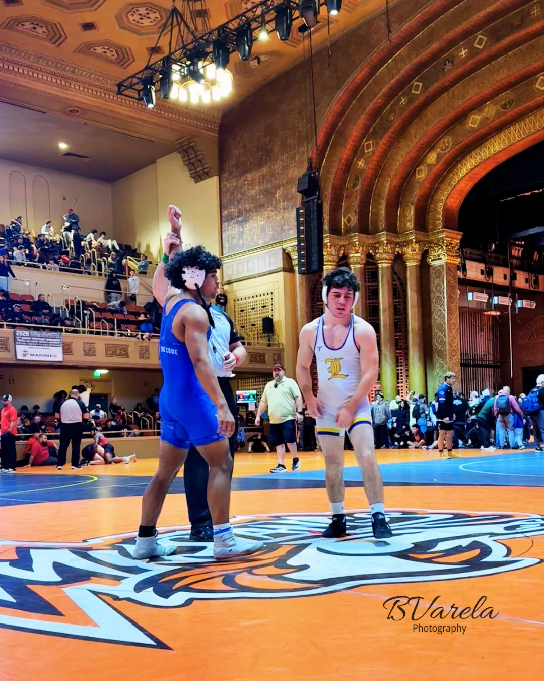 Two wrestlers compete on an orange mat in an ornate indoor arena, with one raising his arm in victory.