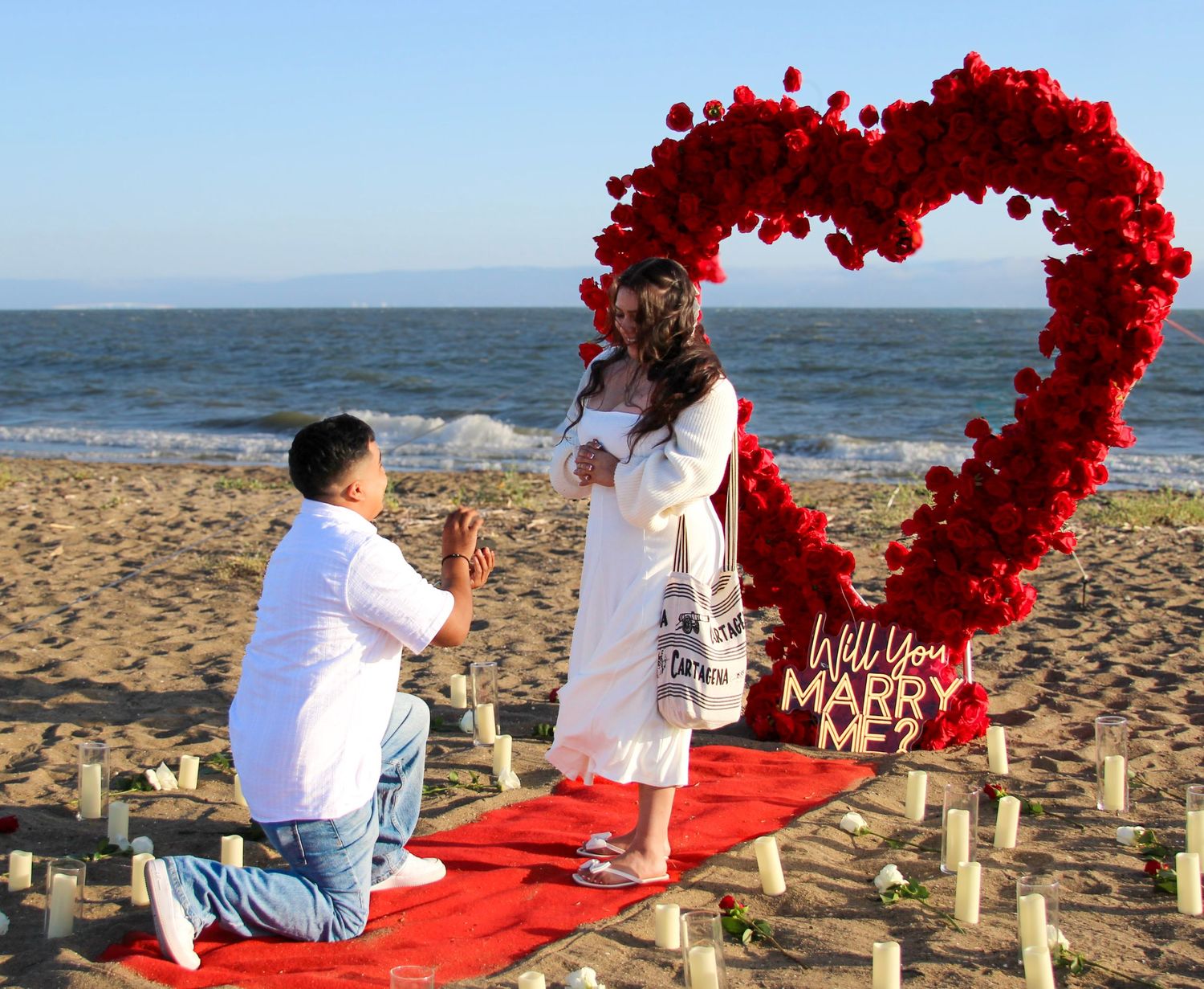 Man proposing on beach with red heart floral arch and Will You Marry Me sign on romantic red carpet setup.
