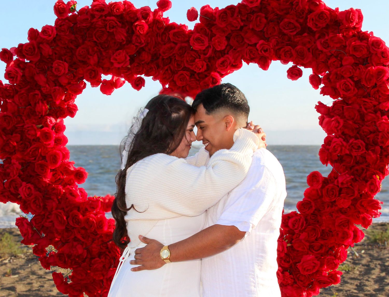 Couple embracing under a red rose heart arch on a beach, dressed in white wedding attire.