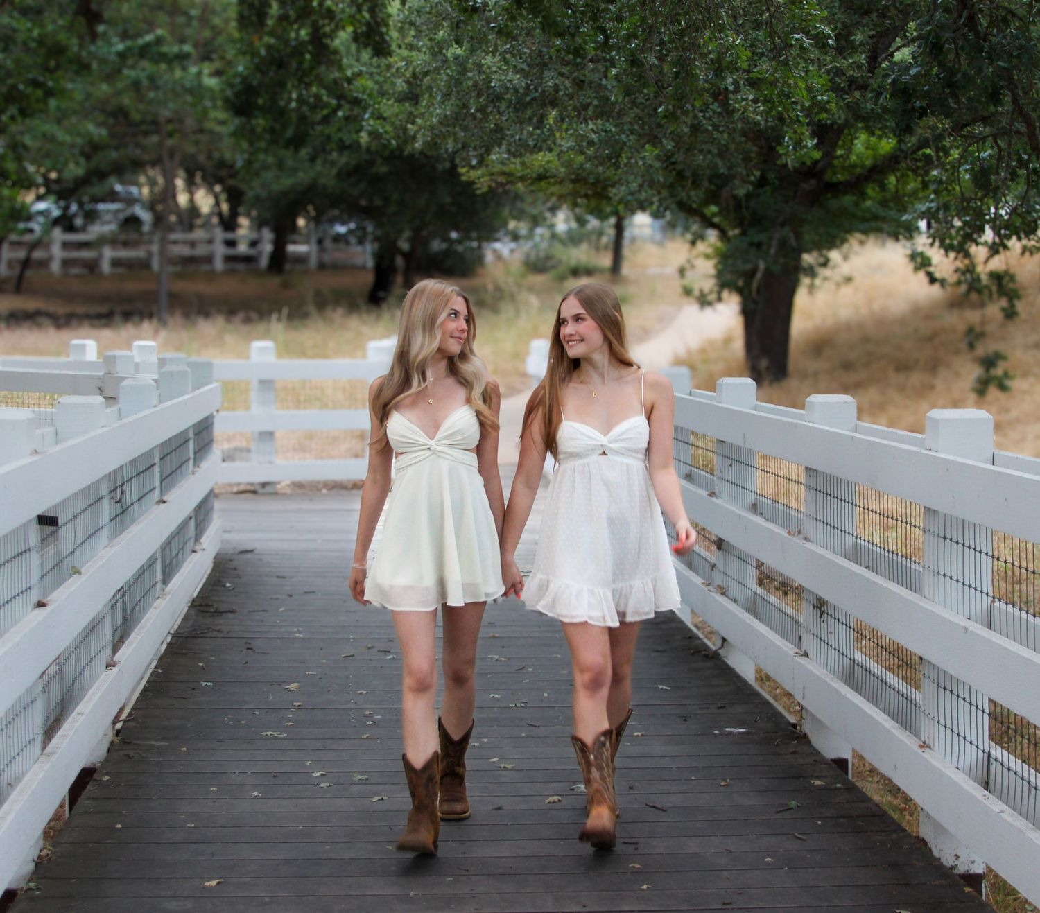 Two young women in matching white mini dresses and cowboy boots walk hand in hand on a wooden bridge.