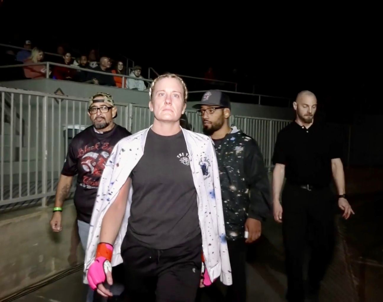 Group of people walking at a nighttime outdoor event, led by a person in a paint-splattered white jacket.