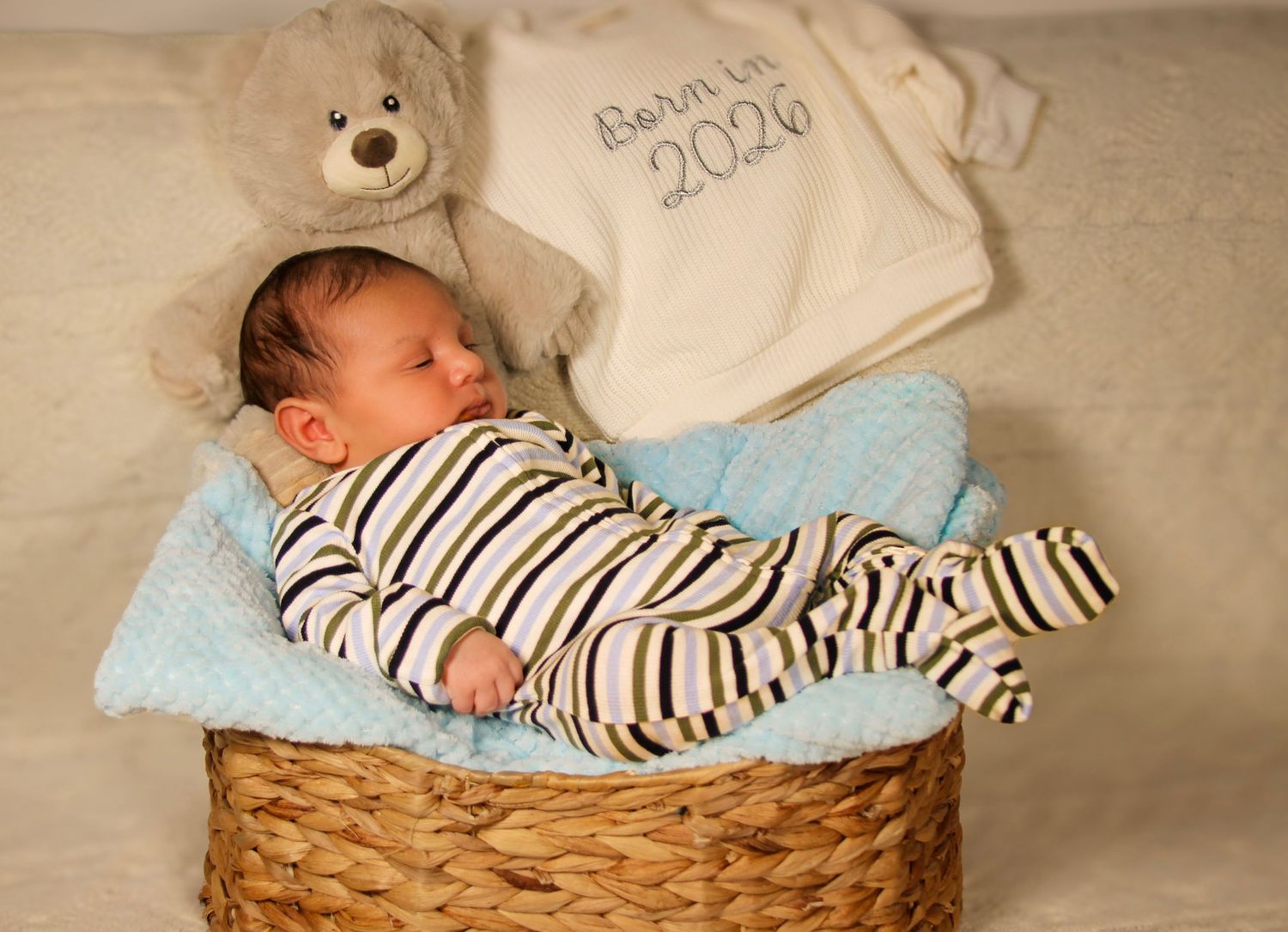 Newborn baby sleeping peacefully in a wicker basket with a teddy bear and Born in 2026 blanket.