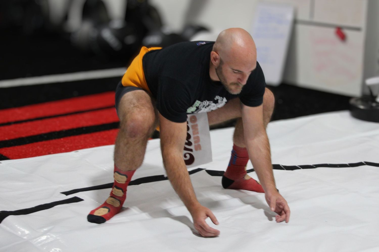 Bald MMA fighter in black shirt and red ankle guards crouches on white mat during combat sports training.