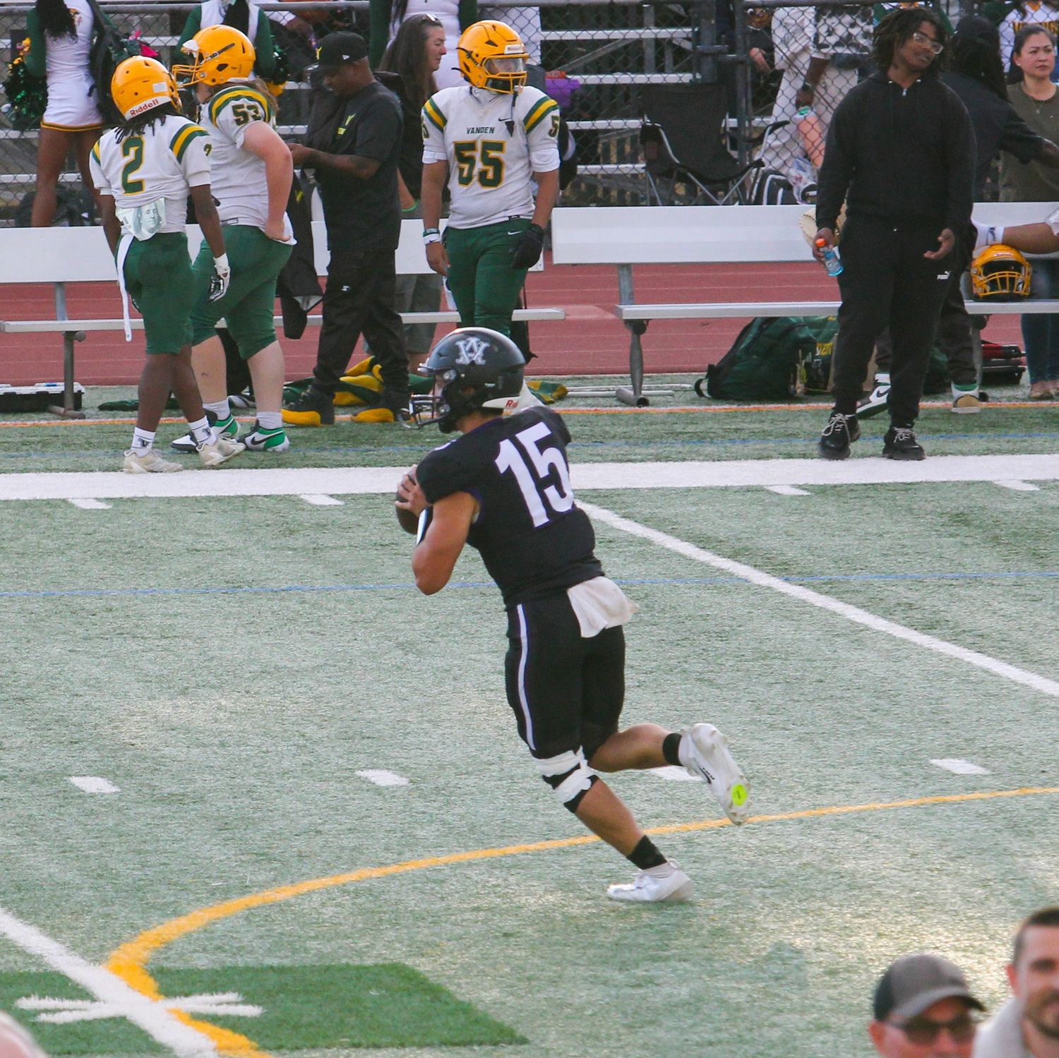 Football player #15 in dark uniform runs on field during game with green and gold team players in background.