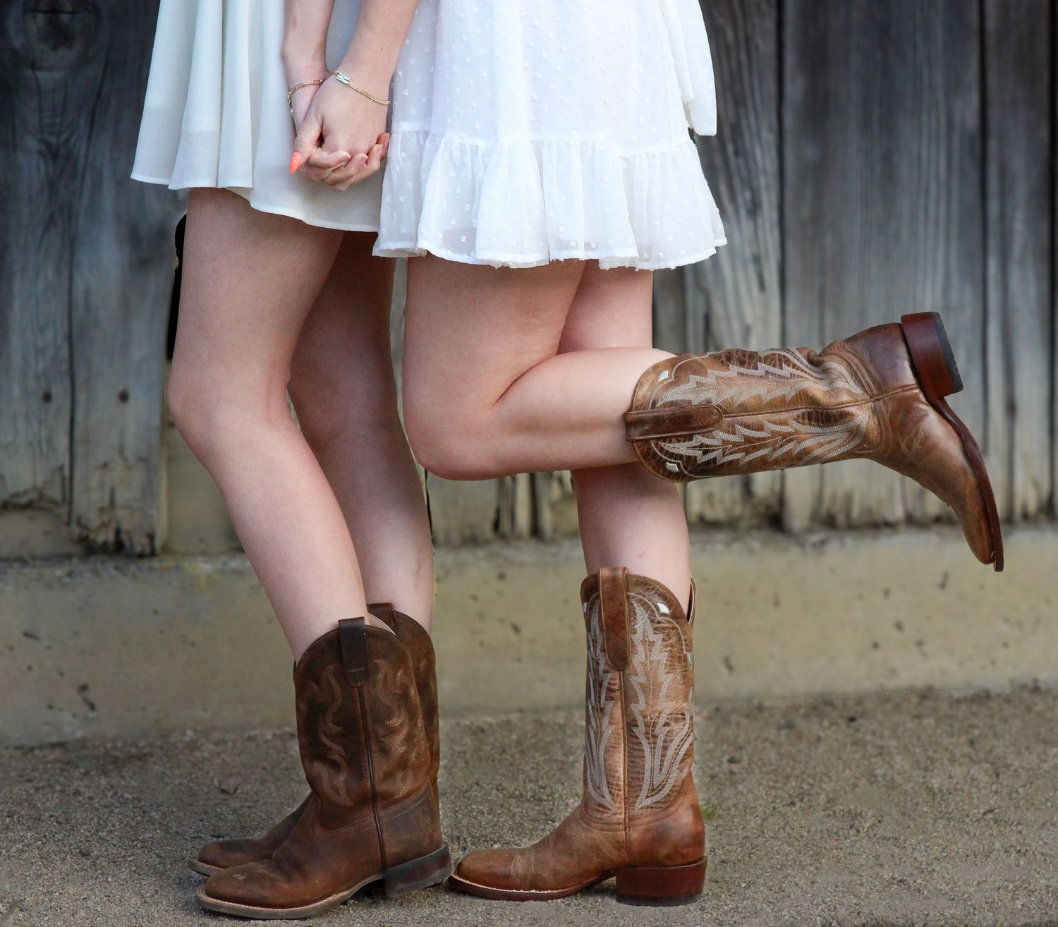 Two women in white dresses and brown cowboy boots pose together near a rustic wooden wall.