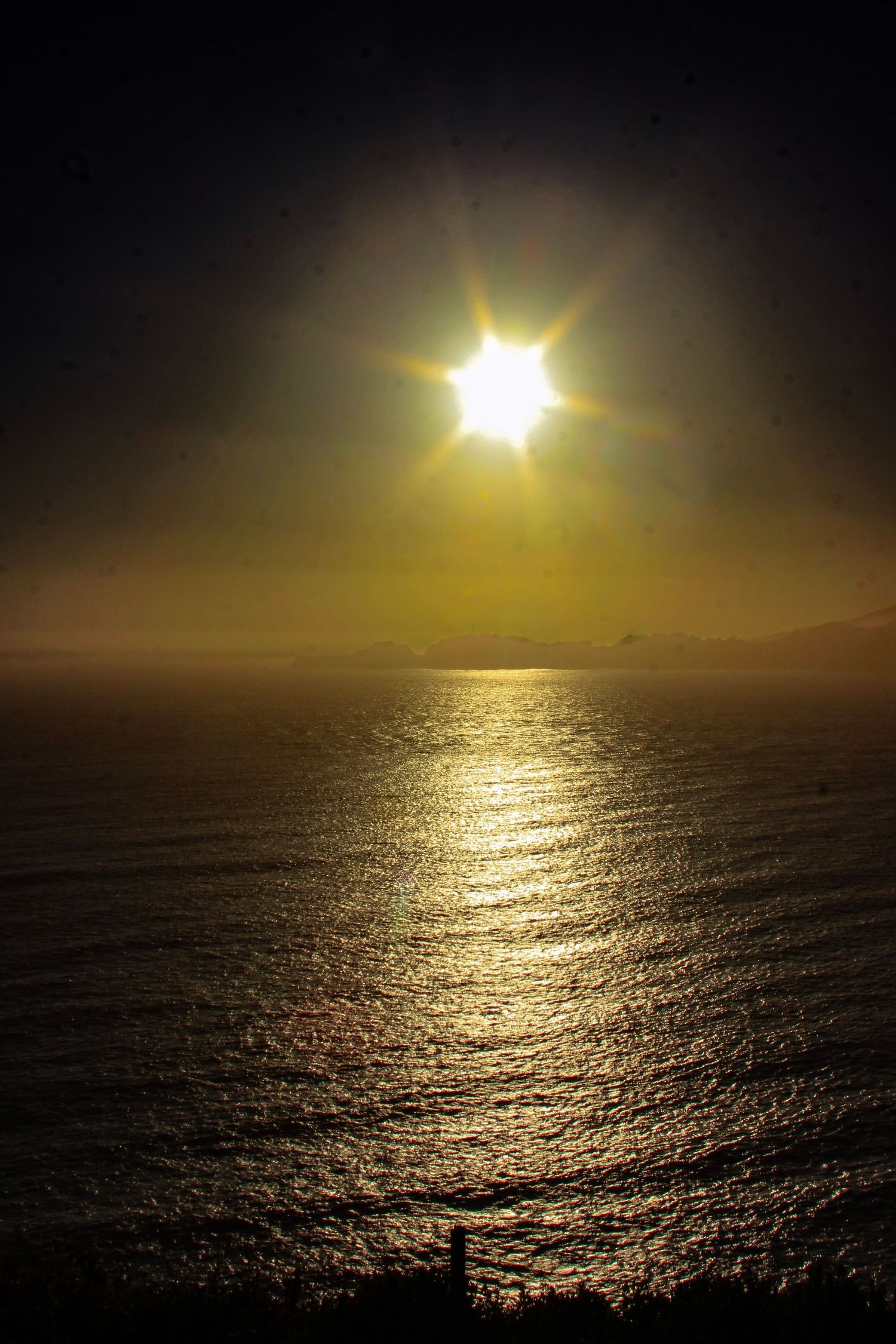 Golden sunset over calm ocean waters with sun rays reflecting on the sea surface against a dark sky.