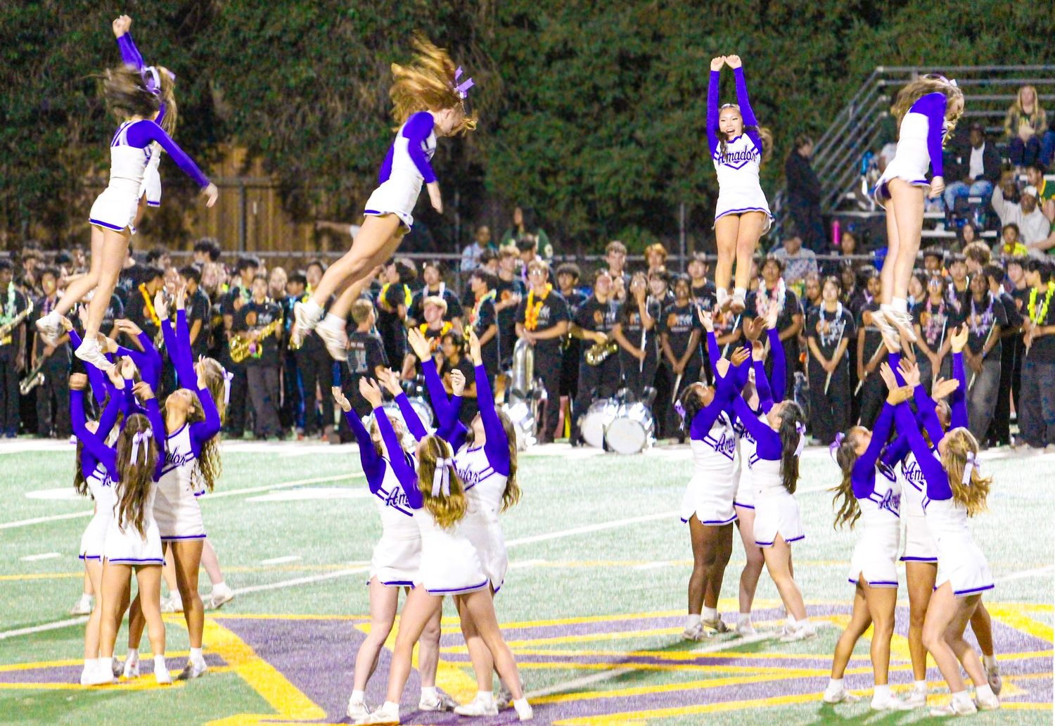 High school cheerleaders in purple and white uniforms perform stunning stunts at a Friday night football game.
