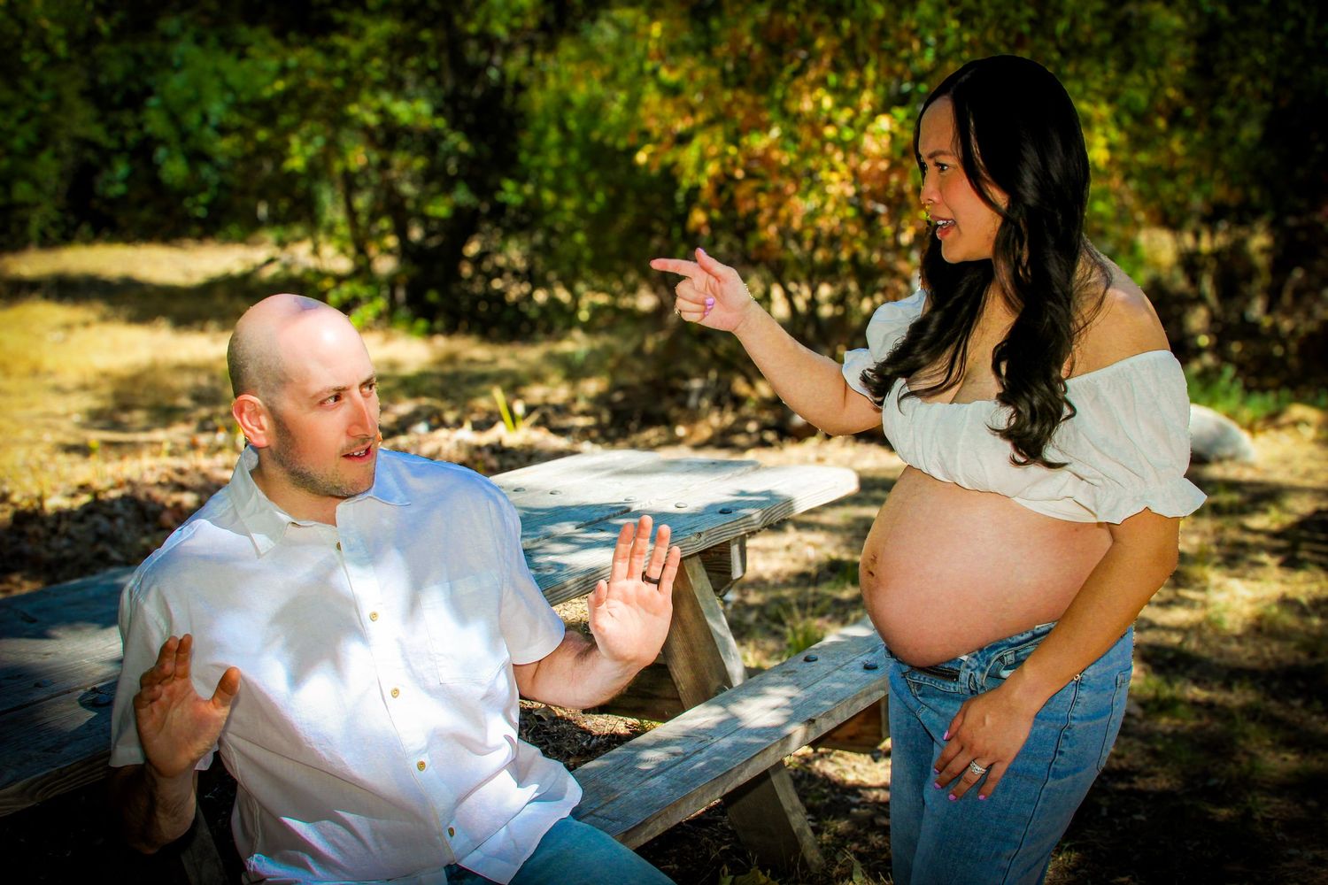 Pregnant woman in white top and jeans pointing at man sitting at picnic table outdoors.