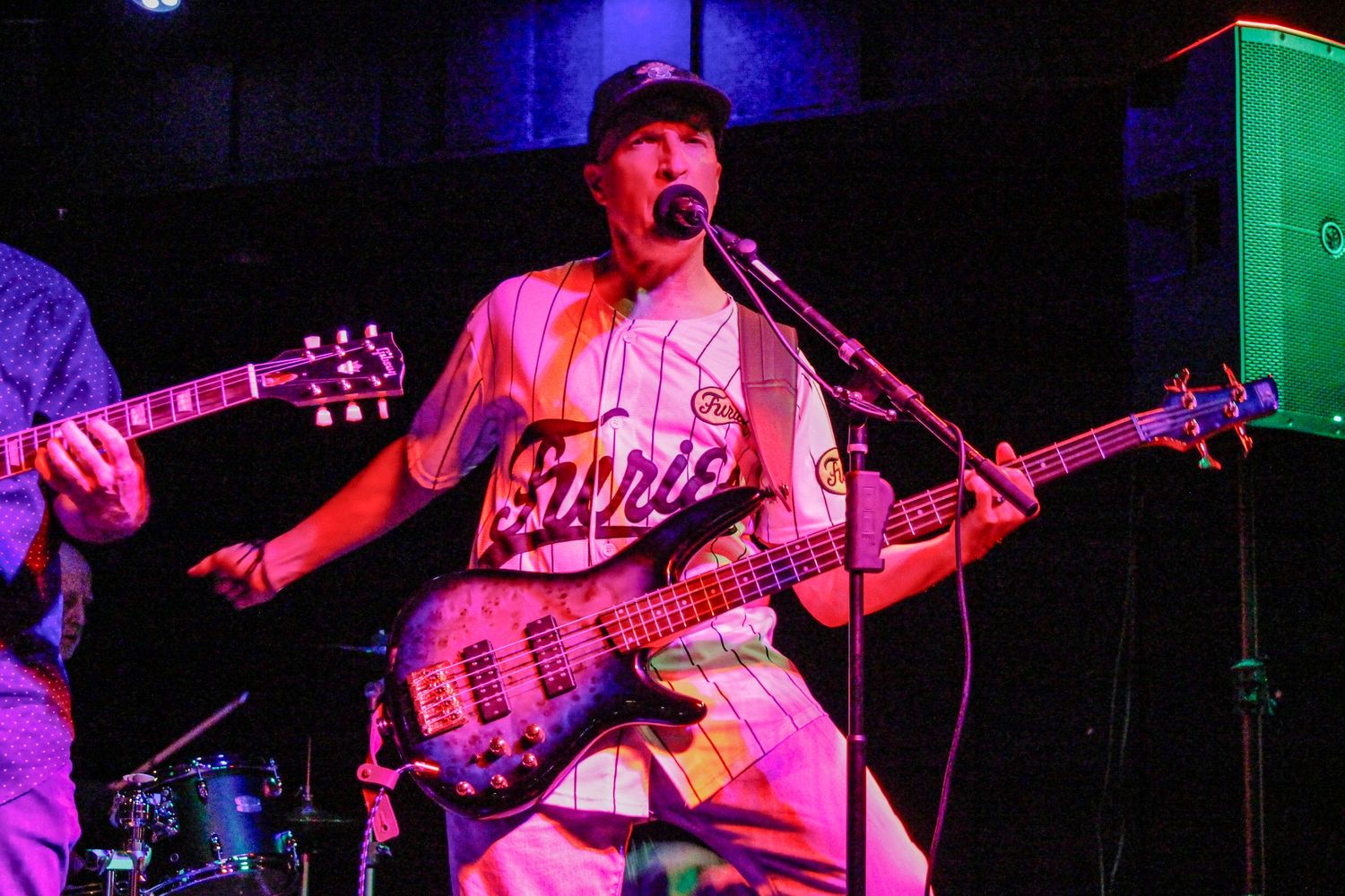 Musician playing bass guitar on stage, wearing a baseball jersey and cap, performing under colorful pink and purple stage lights.