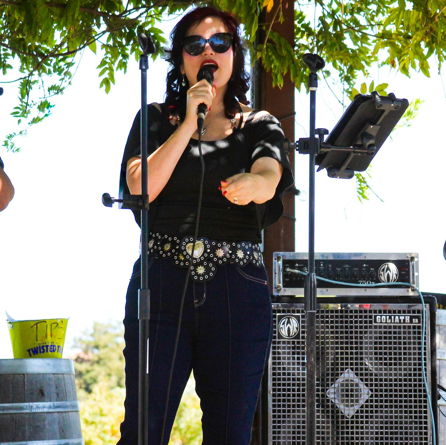 Woman in black outfit and sunglasses singing into microphone on outdoor stage with speaker equipment behind her.