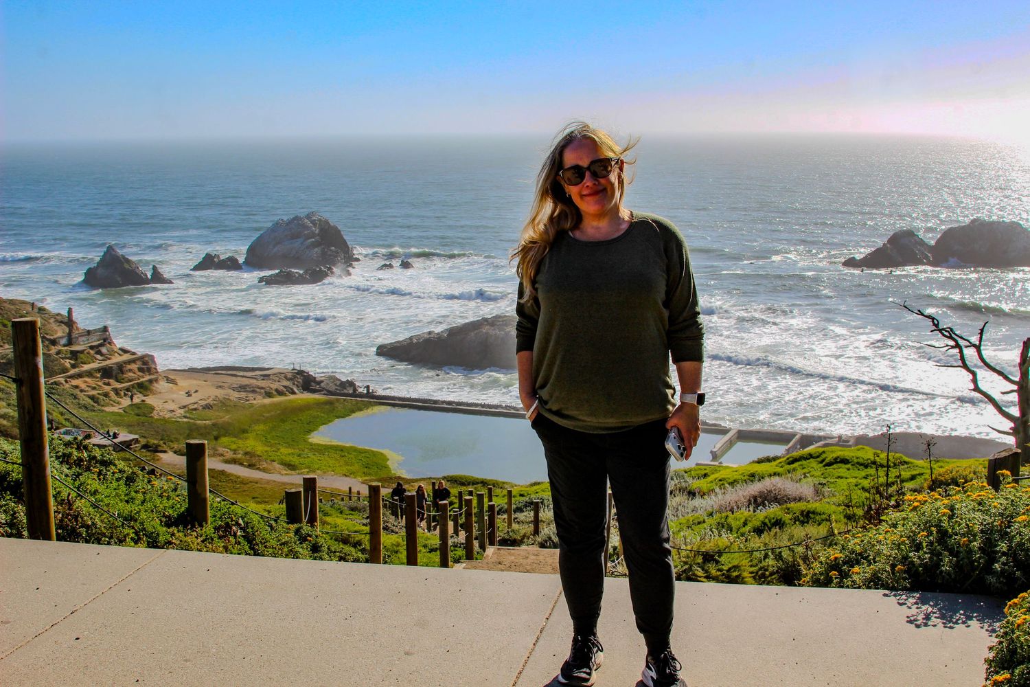 Woman smiling at scenic coastal overlook with rocky ocean shoreline, sea stacks, and blue sky in background.