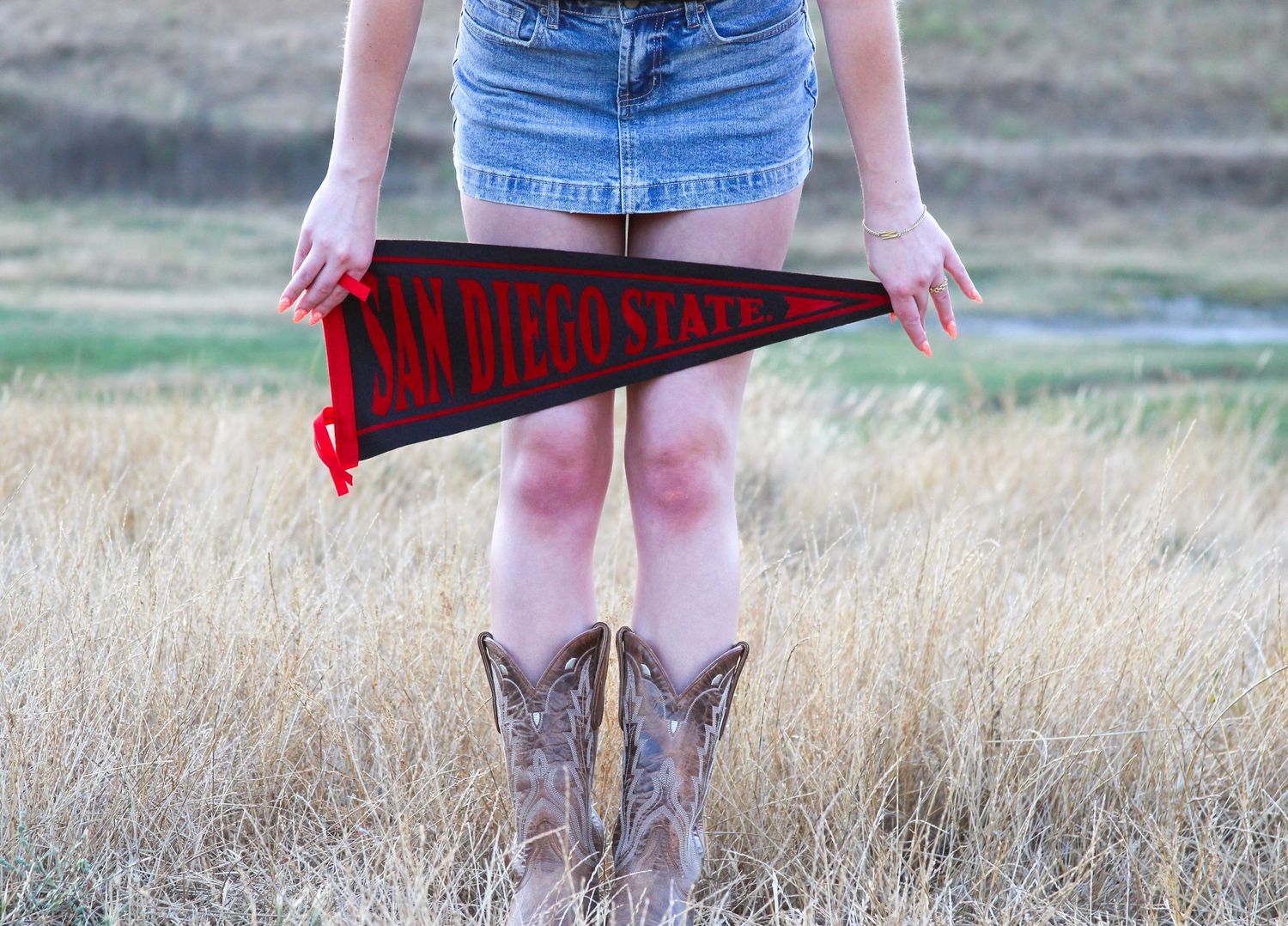 Woman in denim skirt and cowboy boots holding a red San Diego State University pennant flag outdoors.
