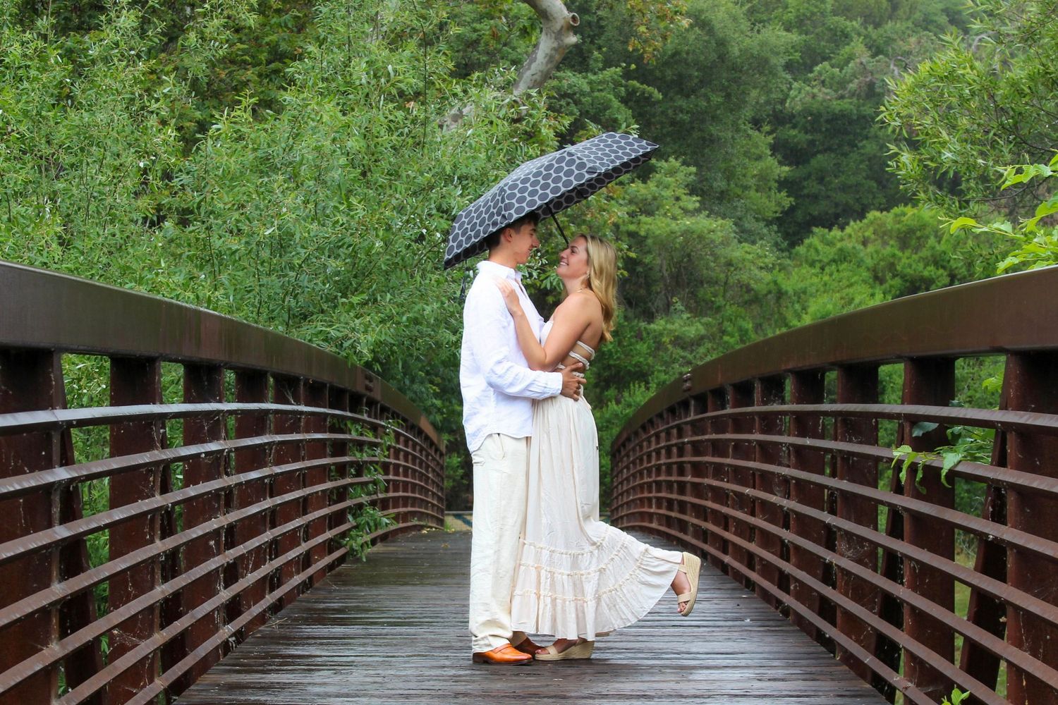 Couple kissing under a polka dot umbrella on a wooden bridge surrounded by lush green trees.