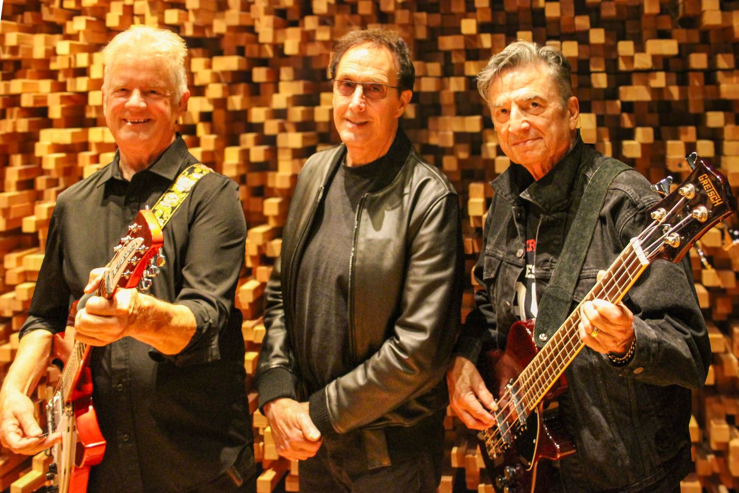 Three men smiling and posing with electric guitars in a recording studio with acoustic foam walls.