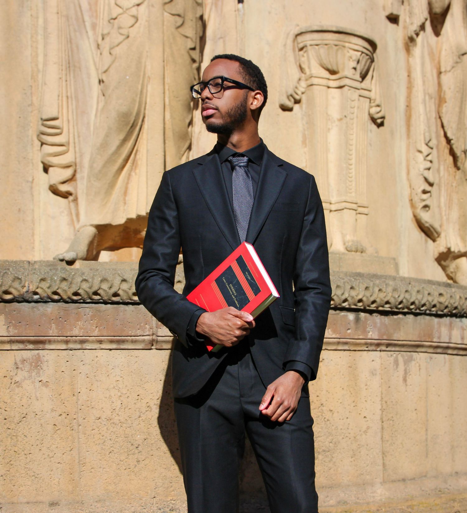 A young man in a black suit holding a red book stands before ornate stone sculptures in sunlight.