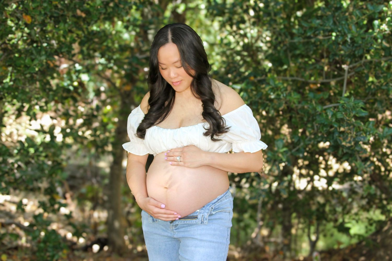 Pregnant woman in white off-shoulder top and jeans cradling her baby bump outdoors among lush green trees.