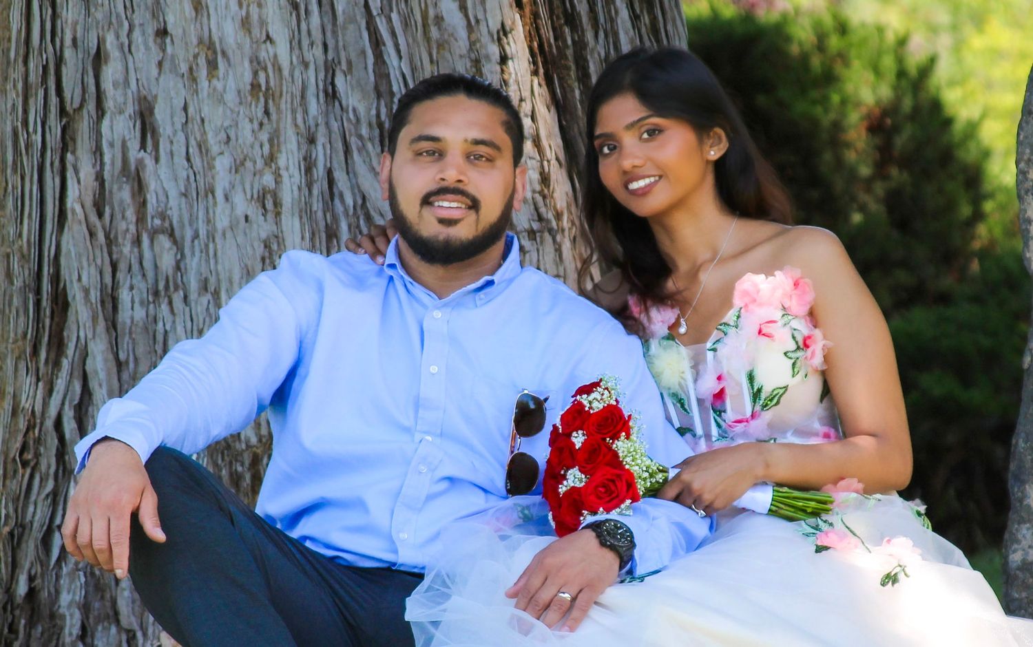 Couple posing by a tree, man in blue shirt, woman in floral white dress holding red roses, outdoor wedding photo.
