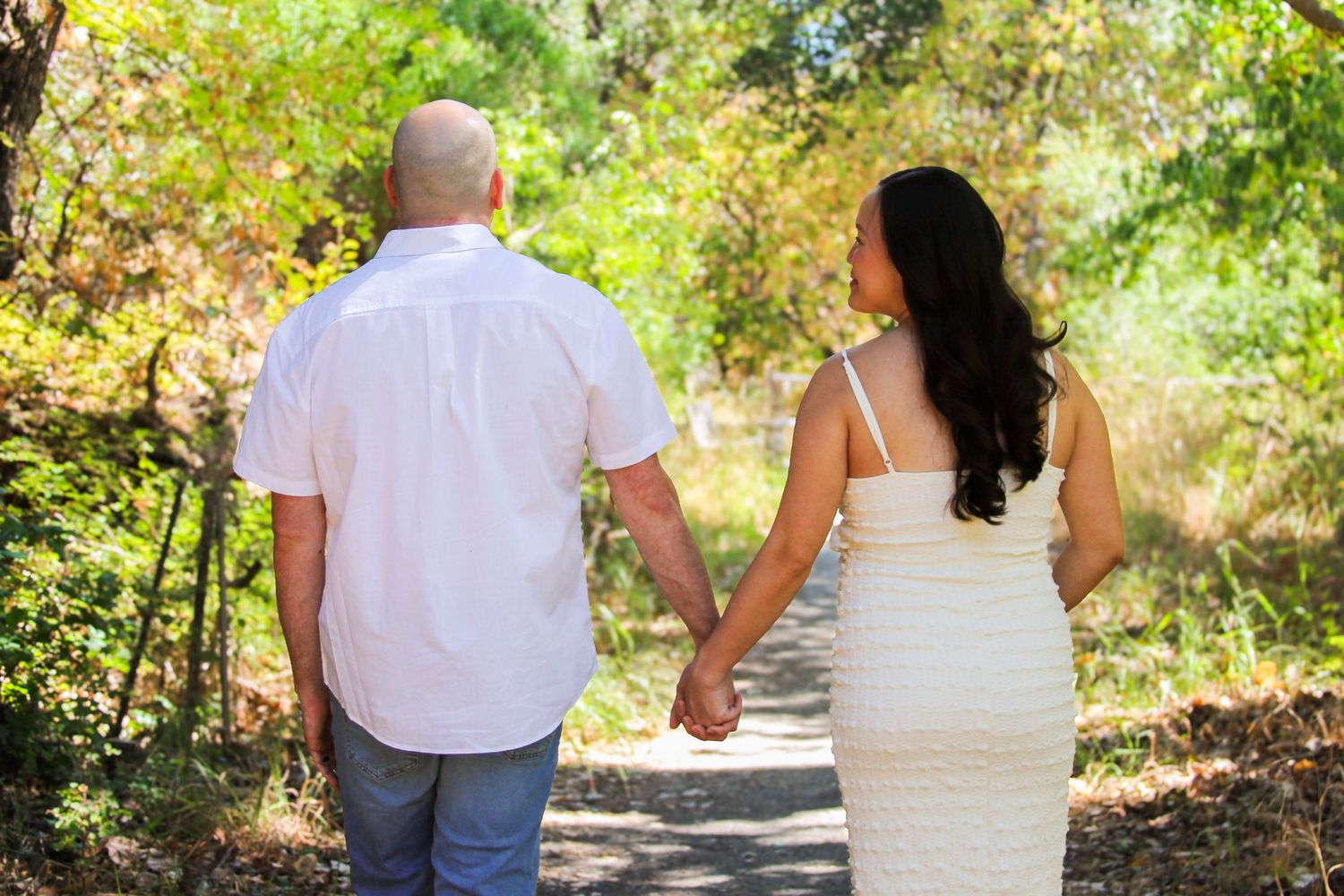 Couple holding hands walking down a tree-lined path outdoors, man in white shirt and woman in white dress.