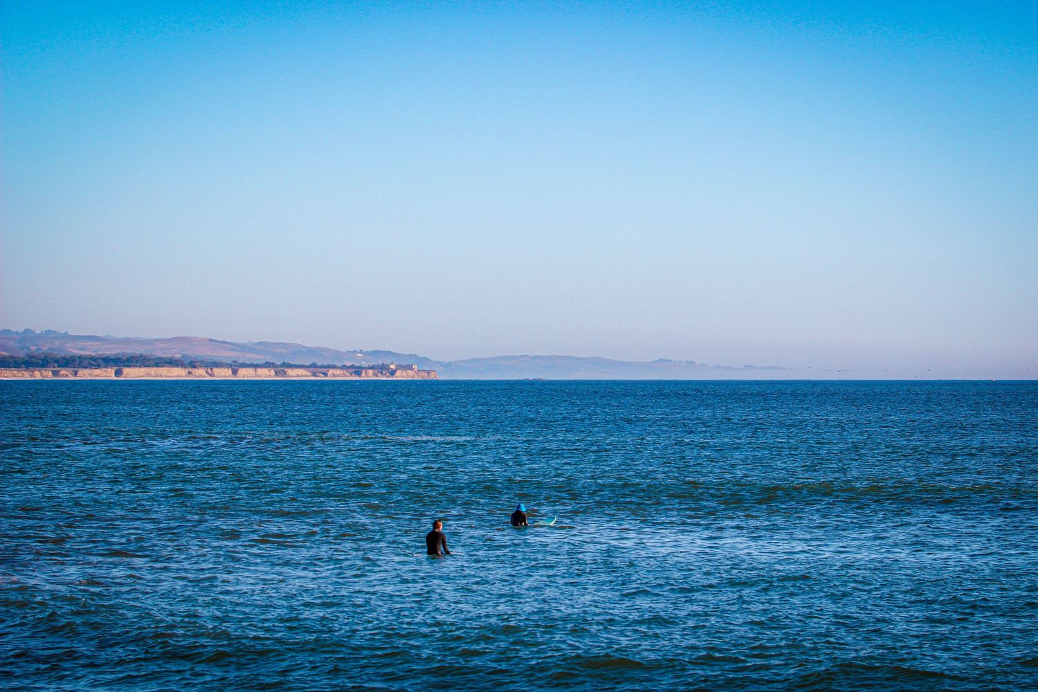 Two surfers wade in calm blue ocean waters with a coastal landscape and clear sky in the background.