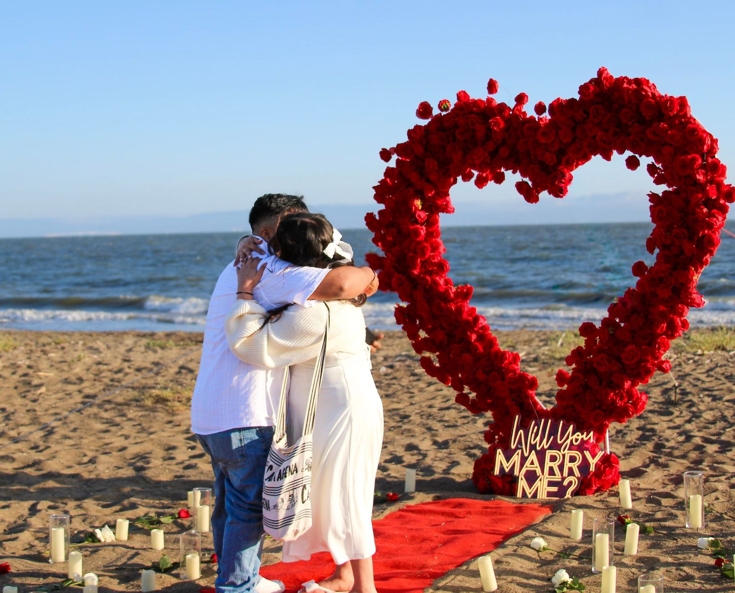 Couple embracing beside a red rose heart arch on a beach with a Will You Marry Me sign at sunset.