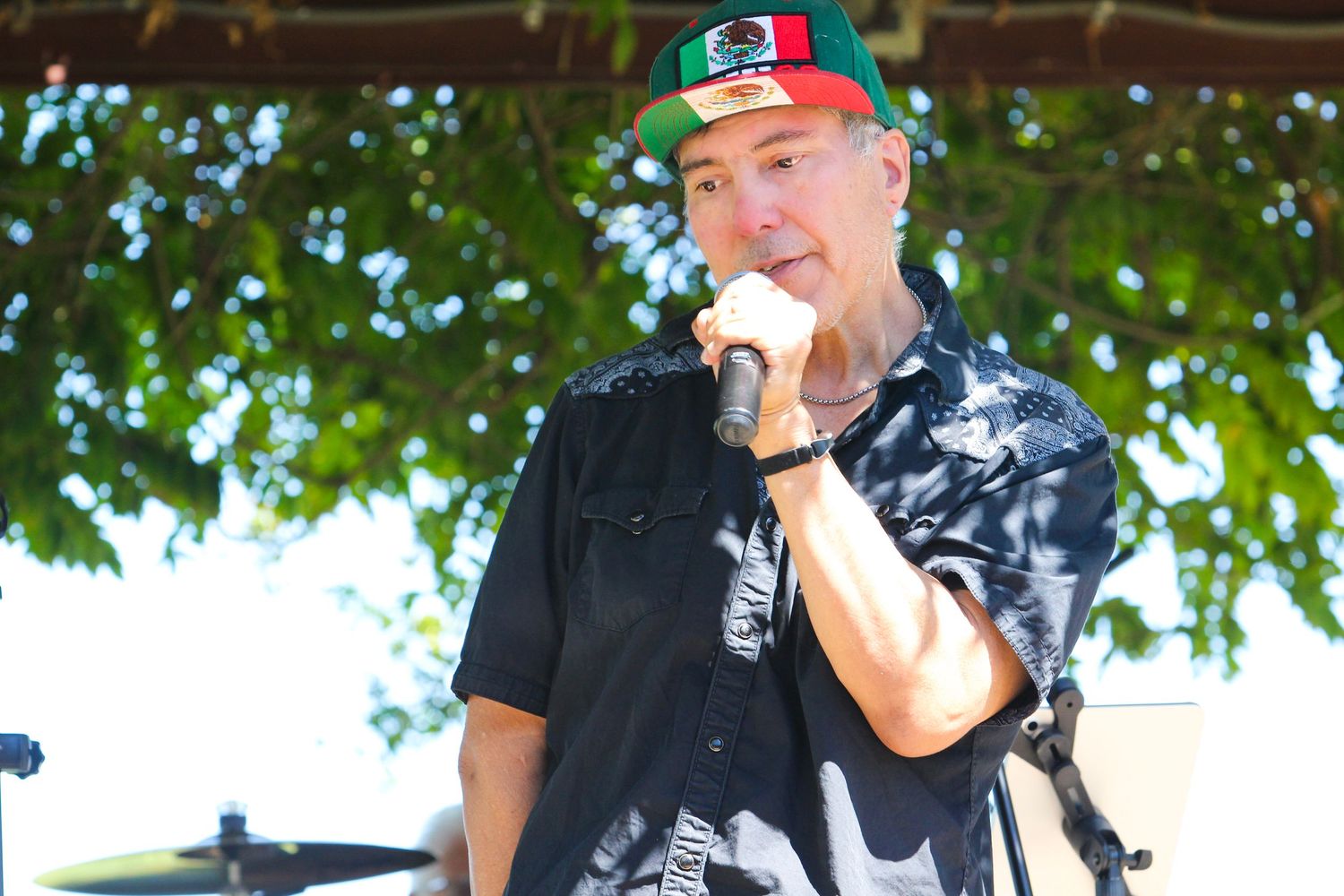 A male singer performs outdoors wearing a colorful Mexican flag cap and black shirt while holding a microphone.