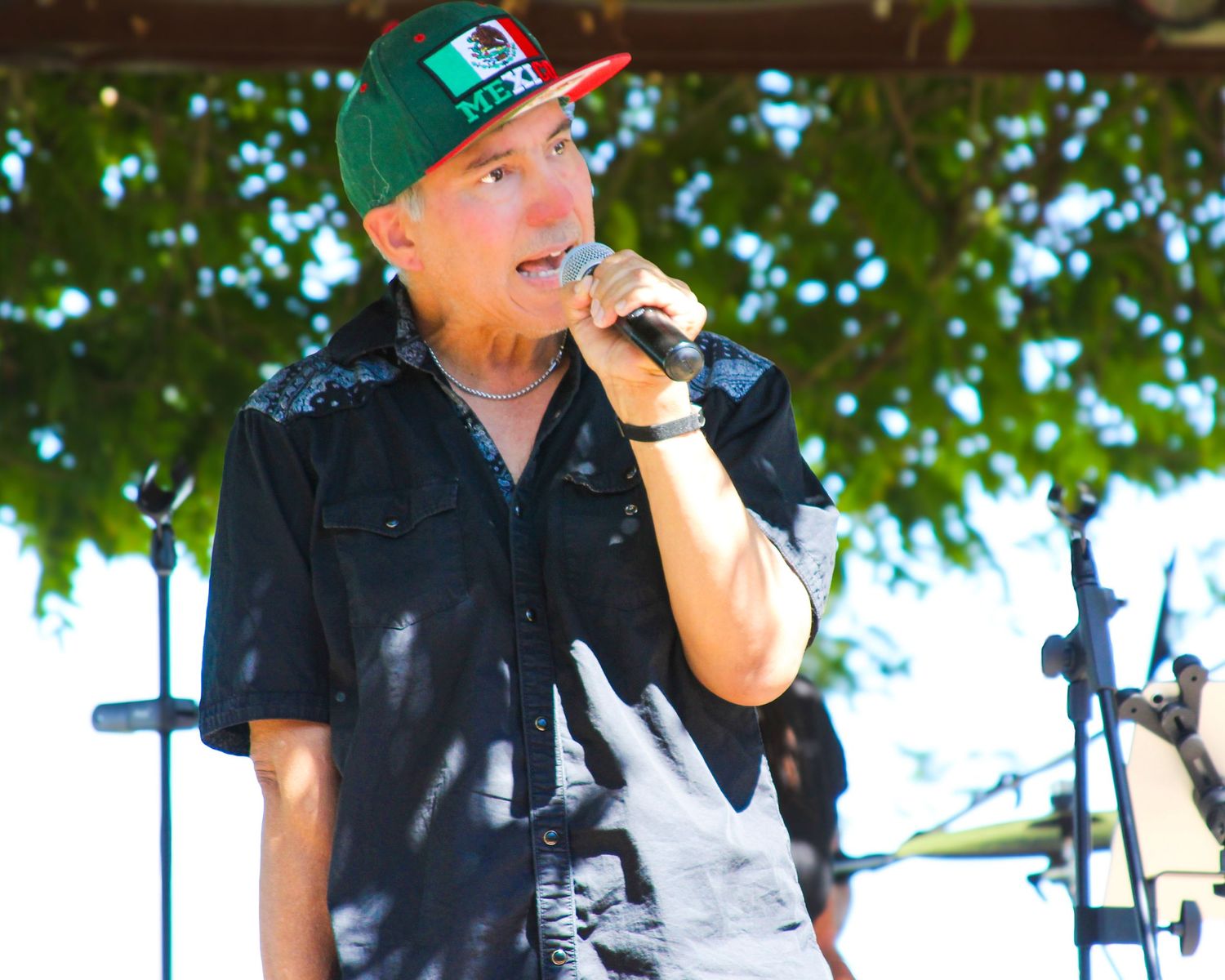 Man singing into microphone outdoors, wearing Mexico flag snapback hat and black shirt at live music event.