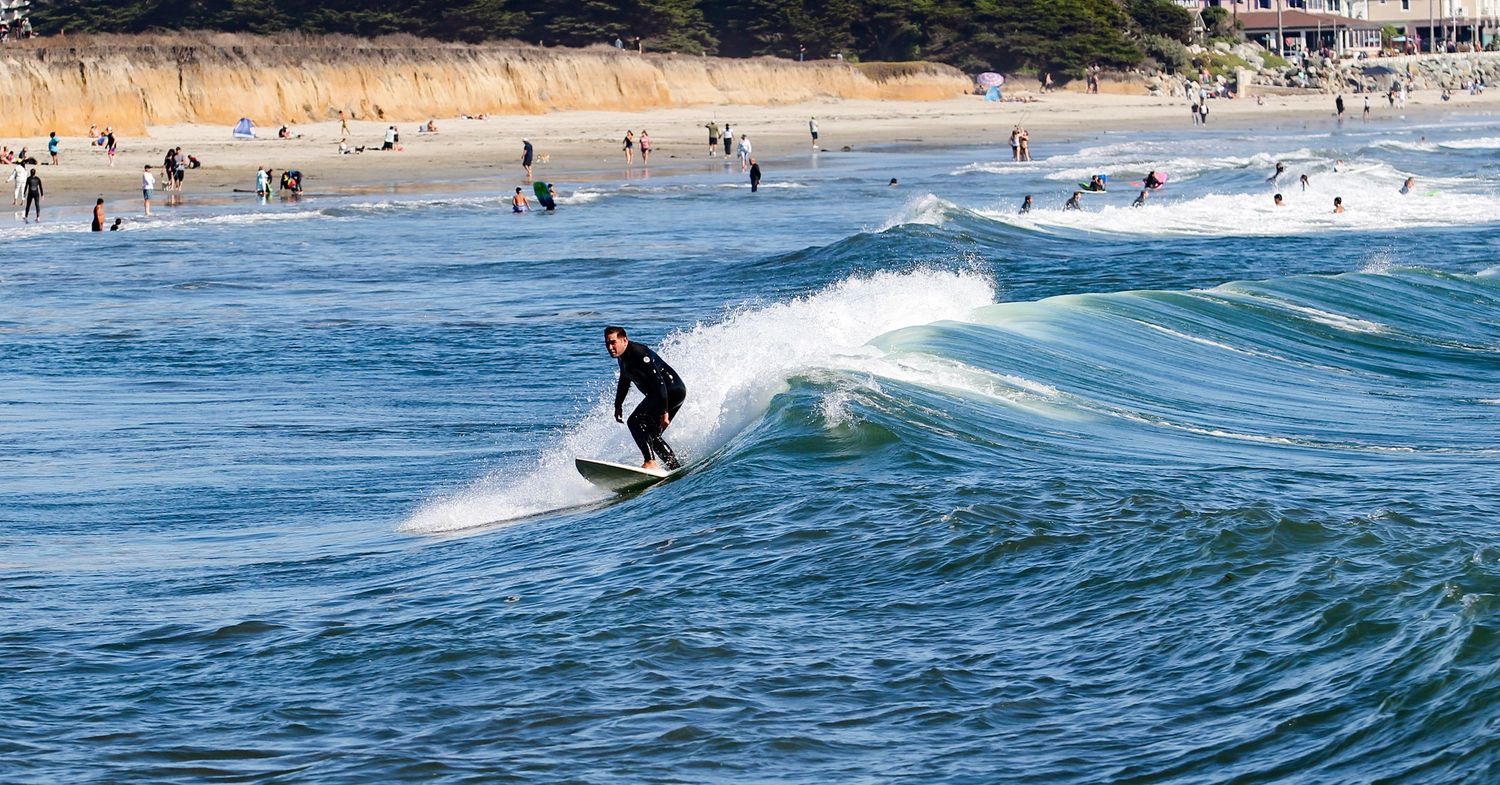 A surfer in a black wetsuit rides a wave at a sunny beach with swimmers and shoreline visible in the background.