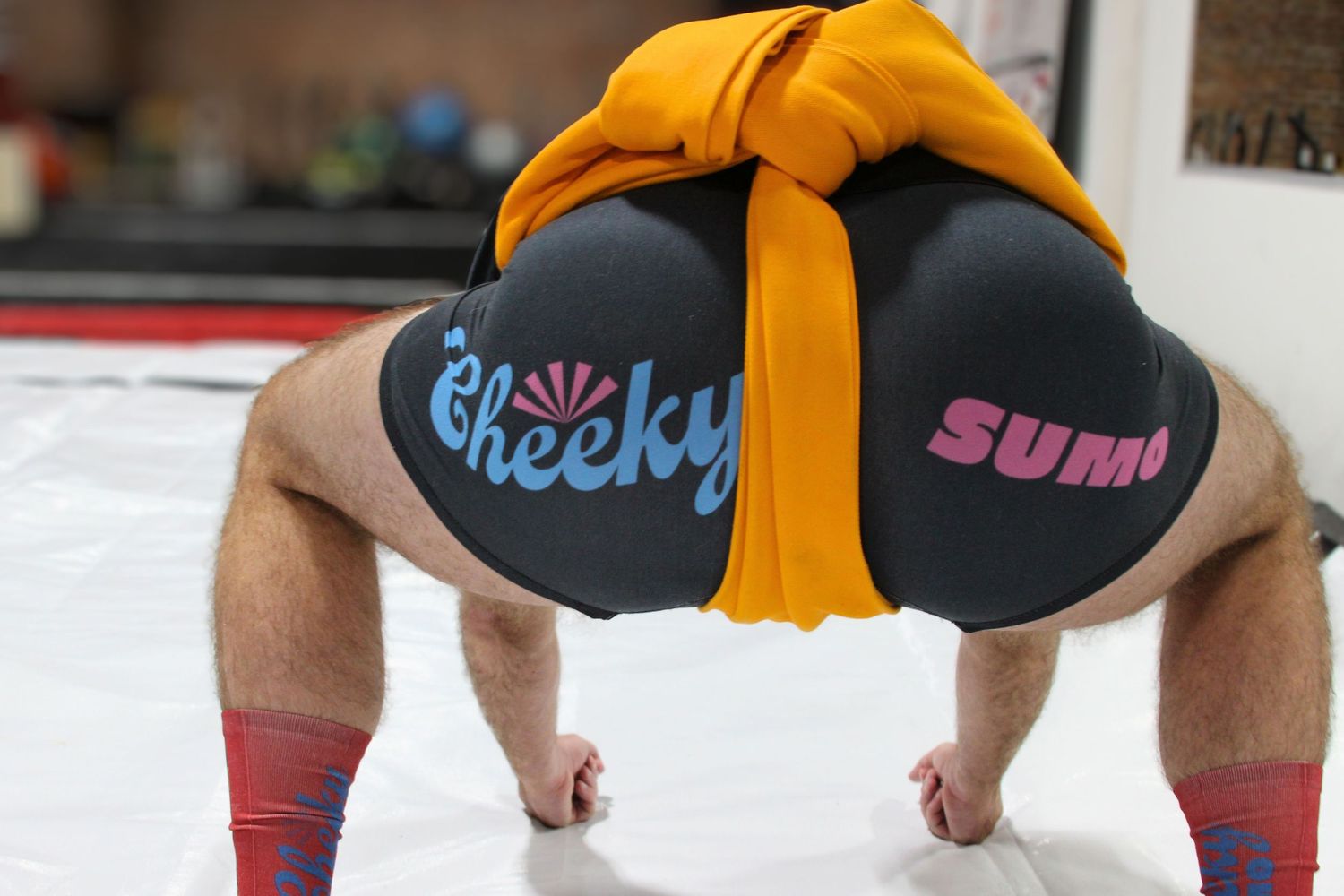 Person bent over wearing Cheeky Sumo branded black shorts with orange sash on a white mat.