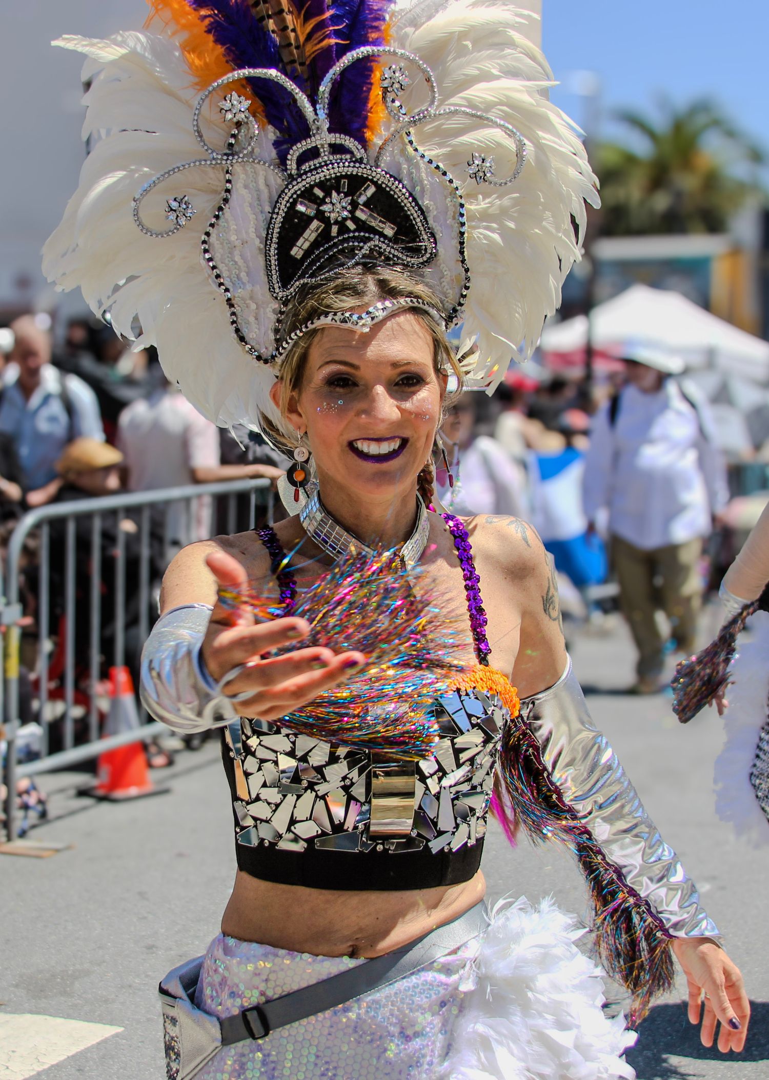 A vibrant carnival dancer in a sequined costume and feathered headdress smiles while performing at an outdoor festival.
