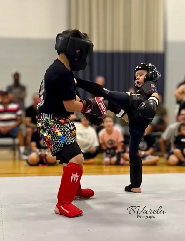 Two young martial artists spar in full protective gear during a youth kickboxing competition in a gymnasium.