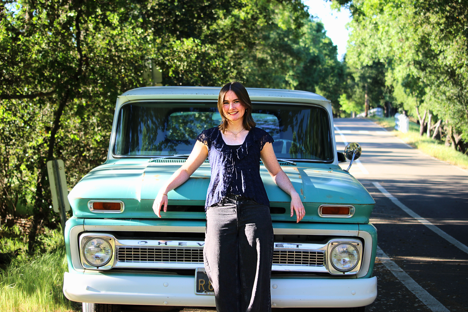 Young woman smiling and leaning against a vintage teal GMC pickup truck on a tree-lined road.