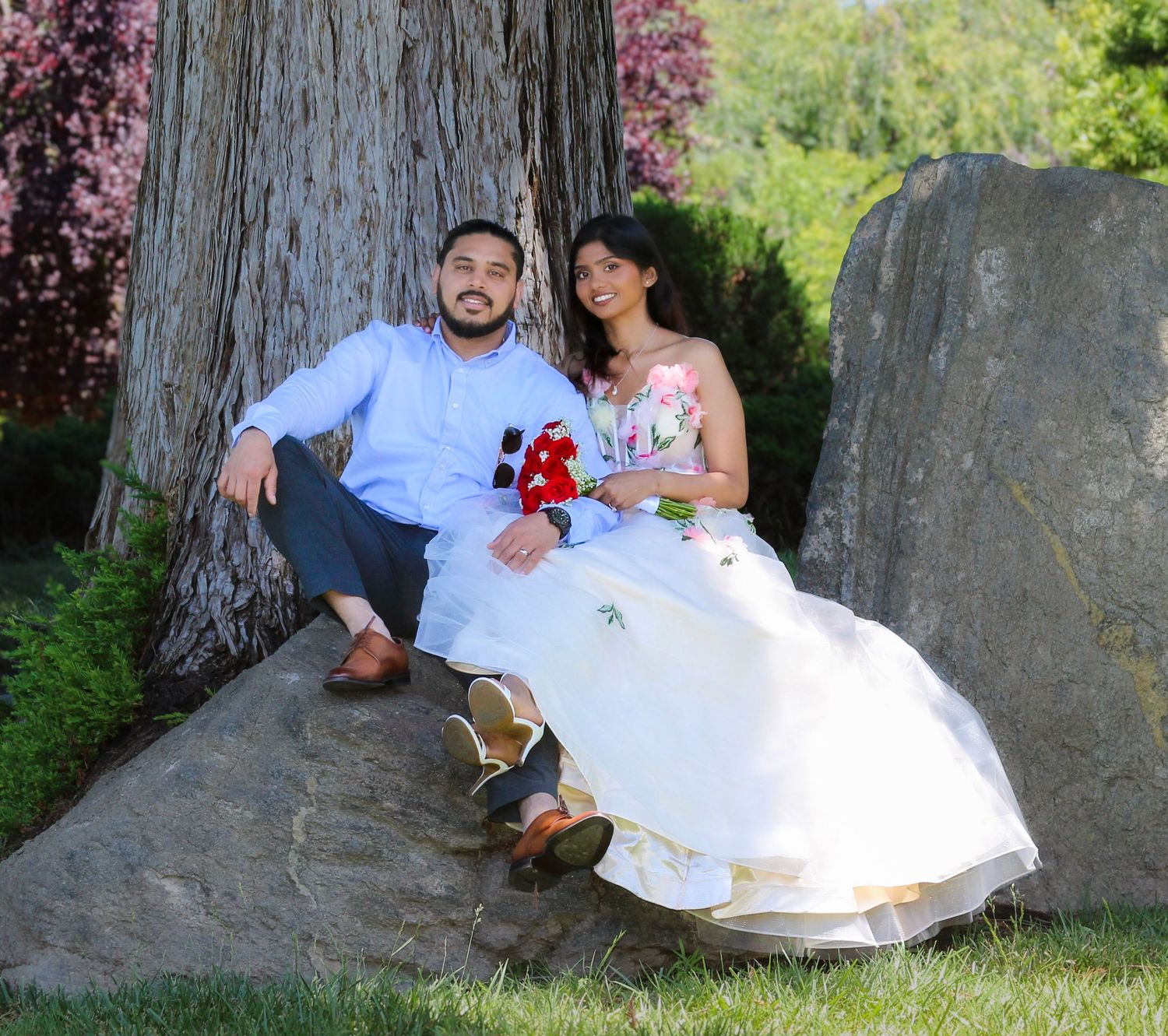 Couple in formal attire sitting on rocks by a large tree in a lush garden, woman in white gown, man in blue shirt.